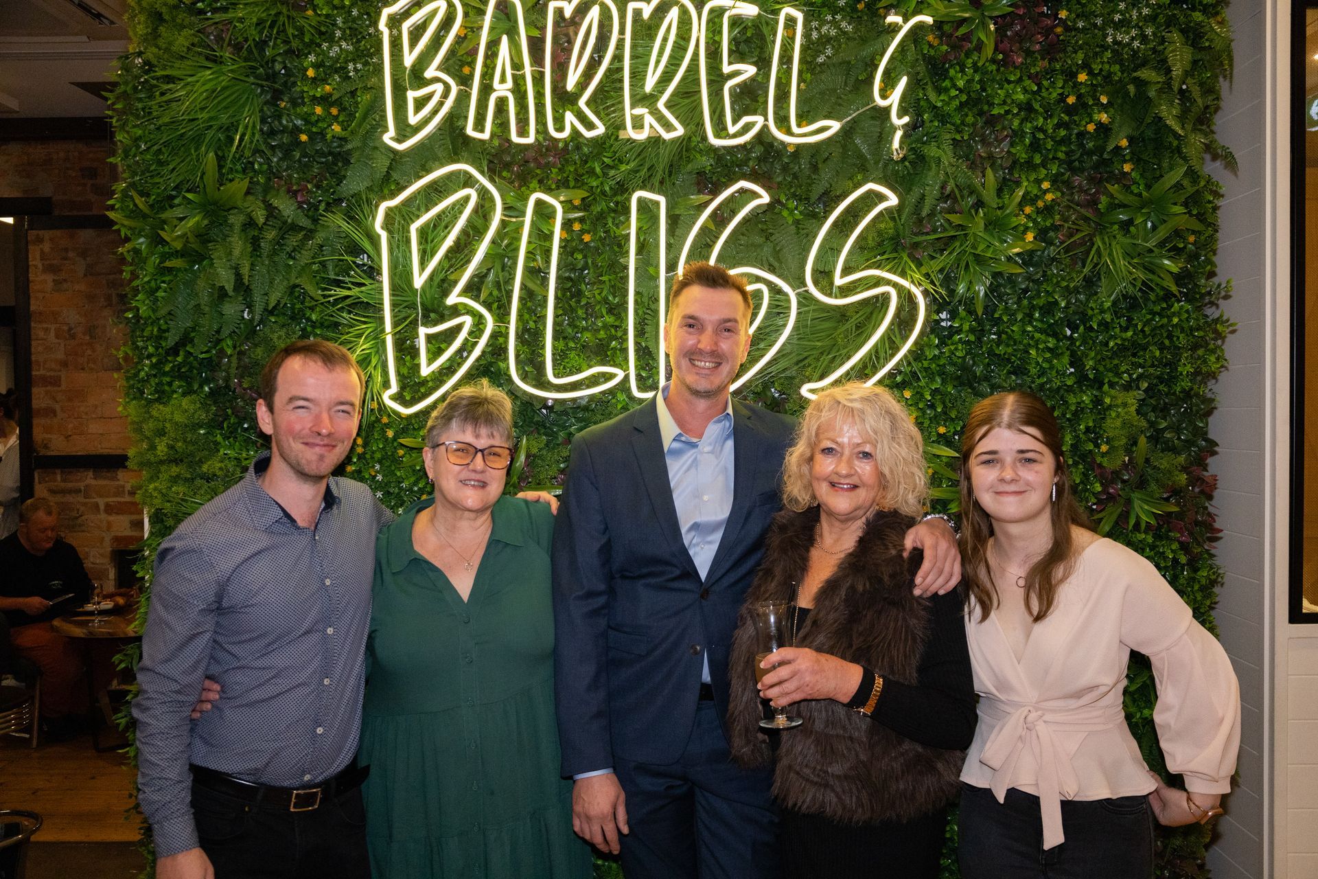 Five people smiling, posing in front of a neon sign that reads 