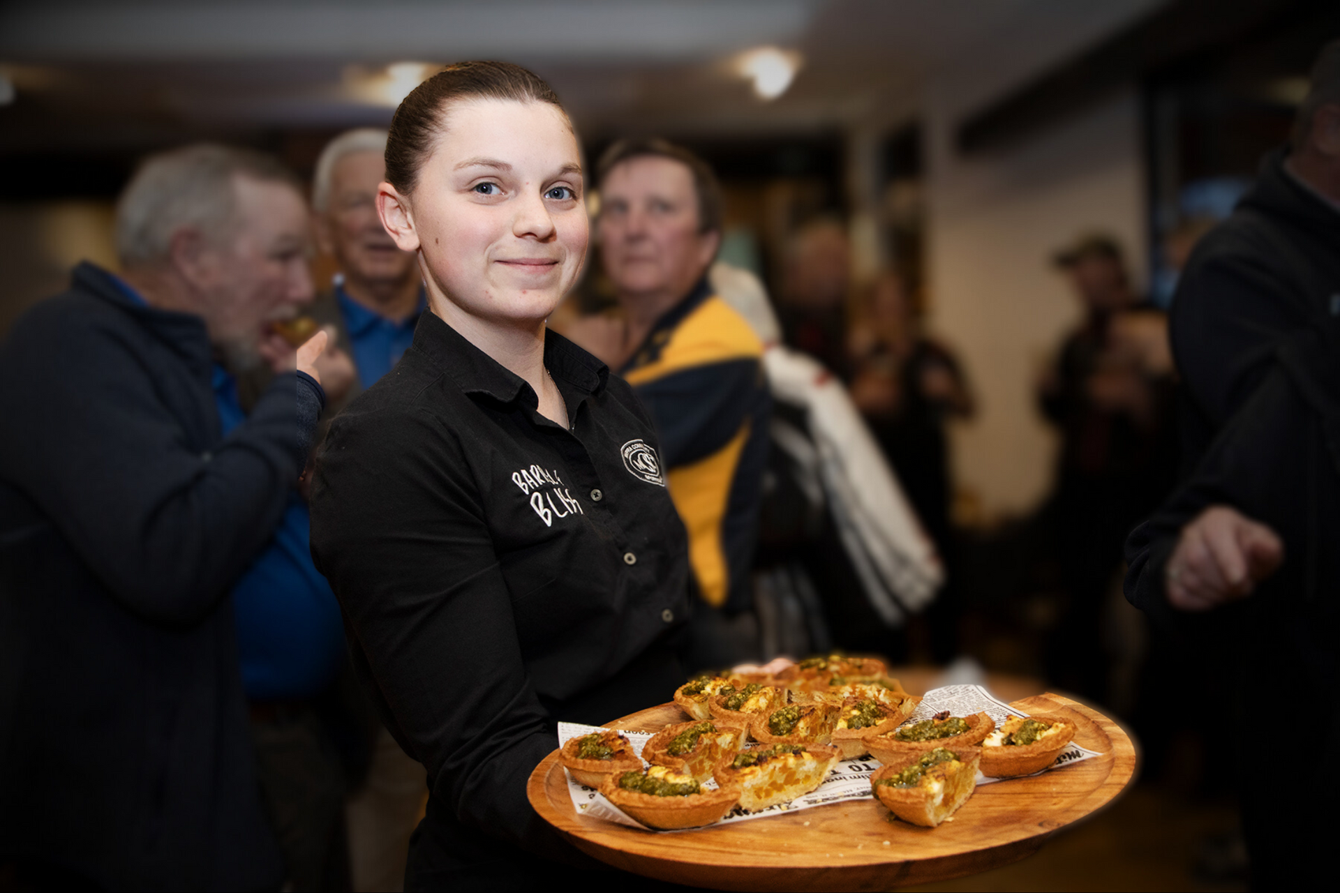 Woman holding tray of food, smiling, with people blurred in the background. Indoor setting, food is light-colored.