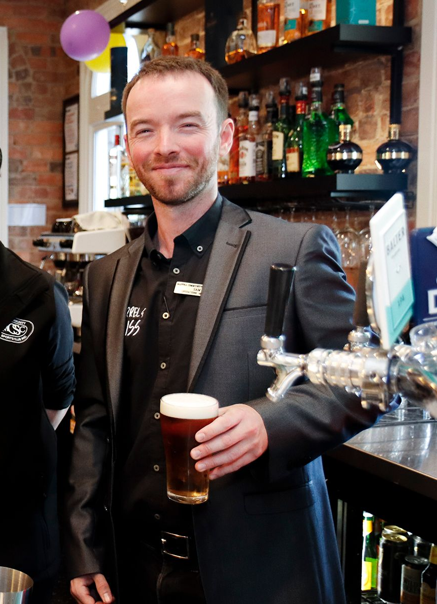 Man in suit holding a beer at a bar; smiles.