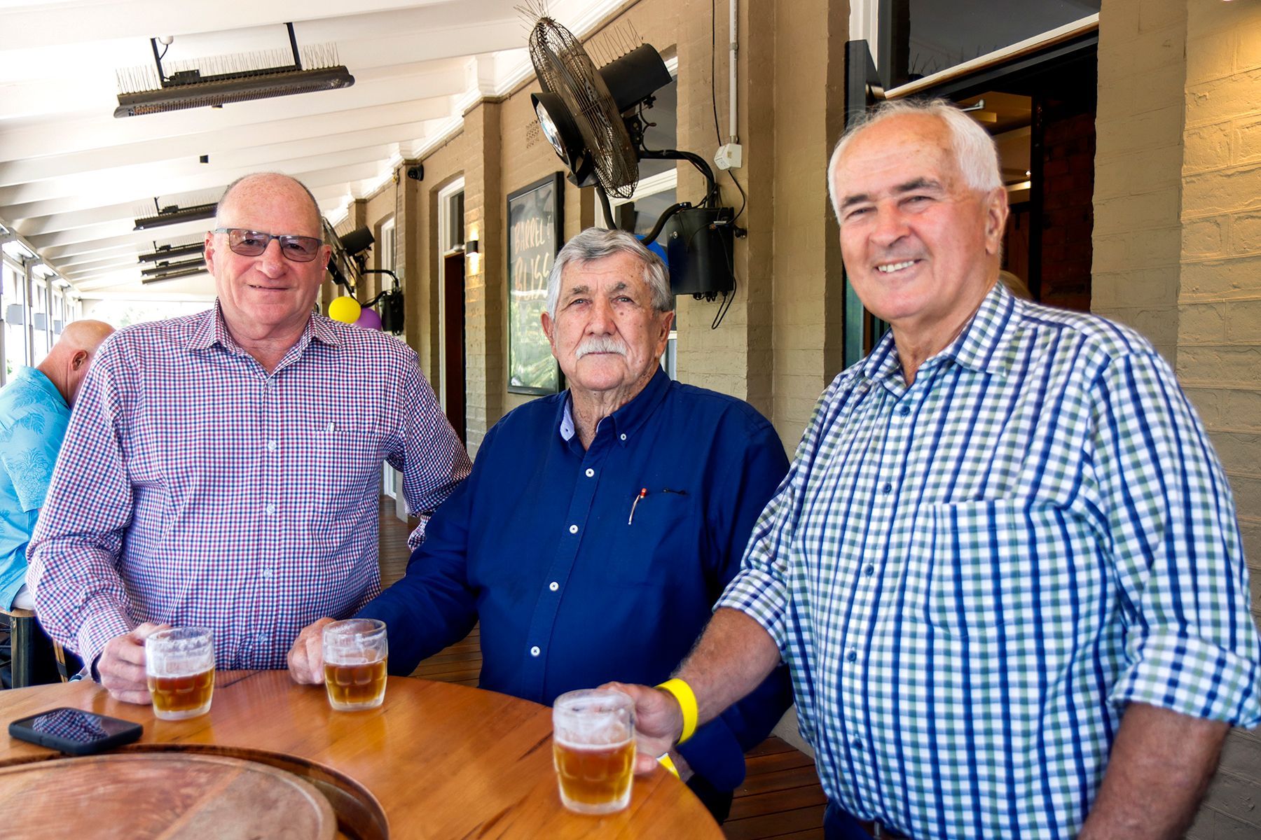 Three men at a bar, holding beers and smiling. Outdoor setting, checkered shirts.
