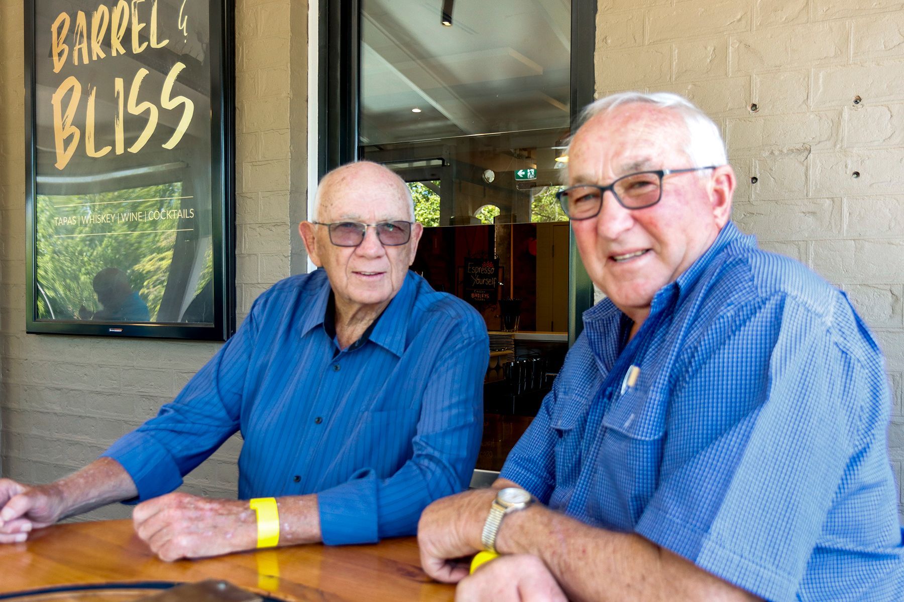 Two elderly men in blue shirts smile, sitting at a table outdoors. 