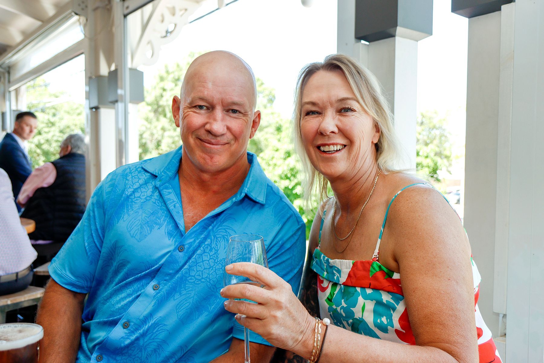 Man and woman smiling, outdoors. Man holds glass of wine, woman wears floral dress.