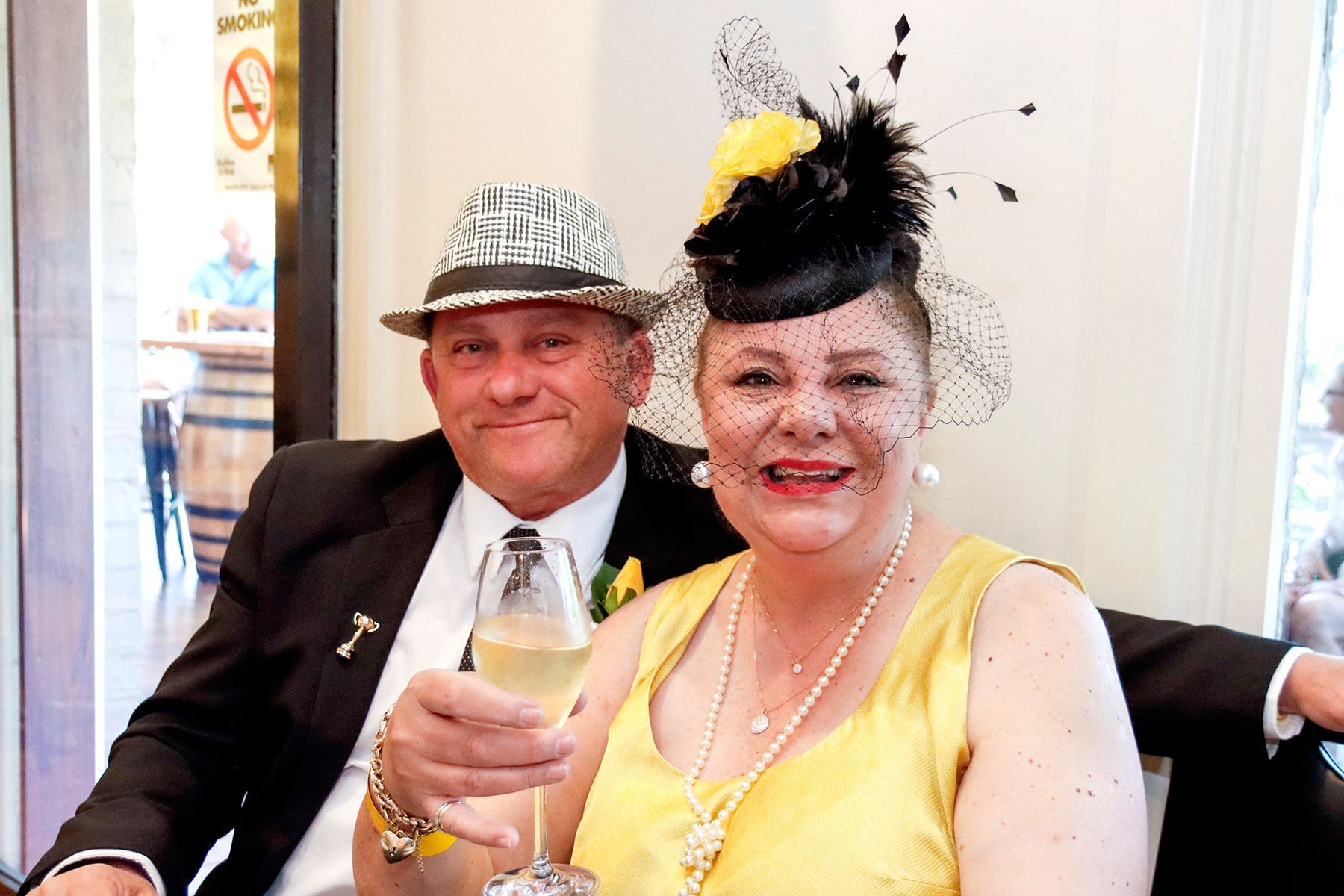 Couple in formal attire, woman holding champagne. Man in hat, woman wearing a fascinator, smiling. Inside setting.