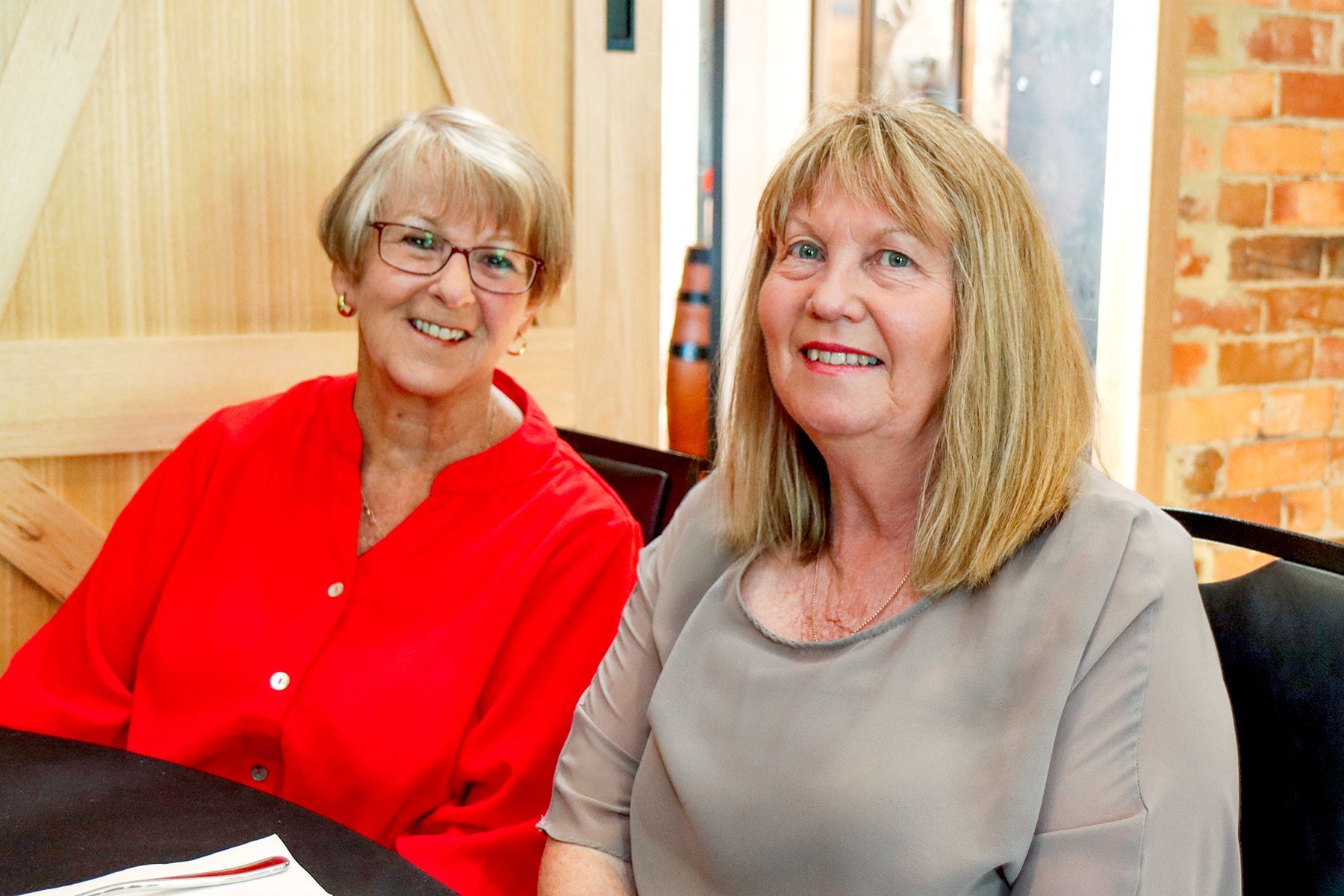 Two smiling women seated at a table; one wearing a red shirt, the other, a beige shirt; light-colored setting.