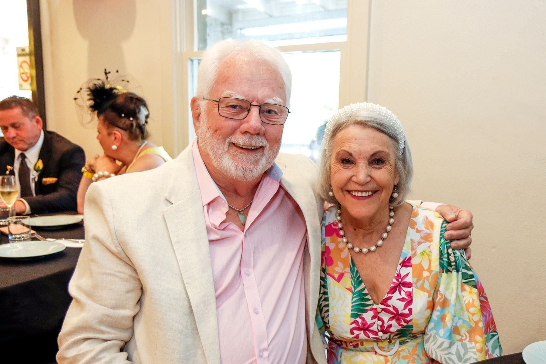 An older couple smiling at a table. The man has a white beard and tan blazer; the woman a floral dress.