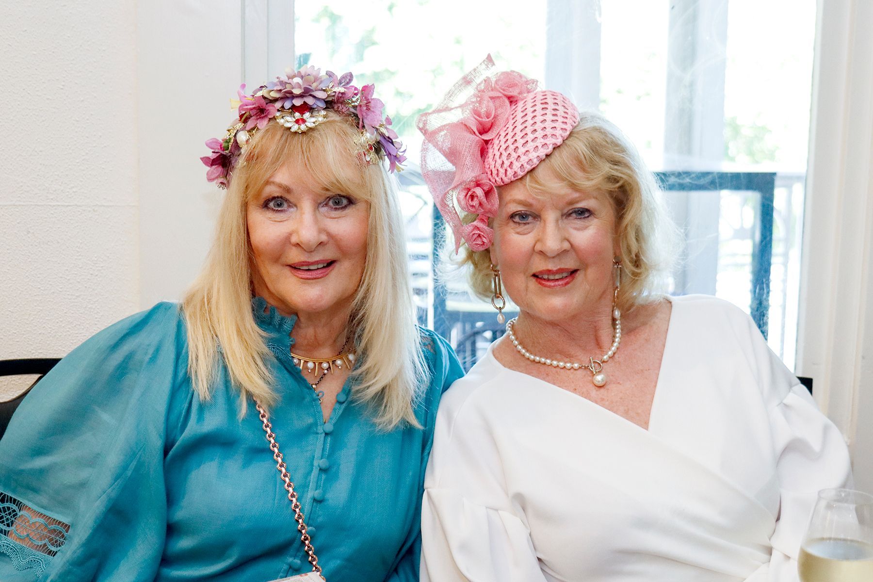 Two older women, blond hair, wearing fancy hats. One in blue, one in white, smiling.