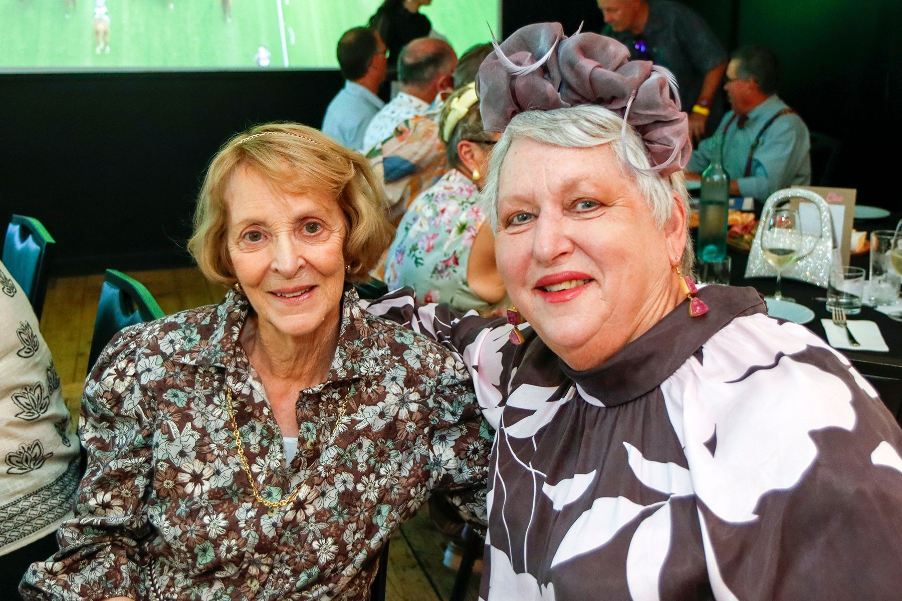 Two smiling women at a seated event. One wears a flowered hat; both are indoors at a table.