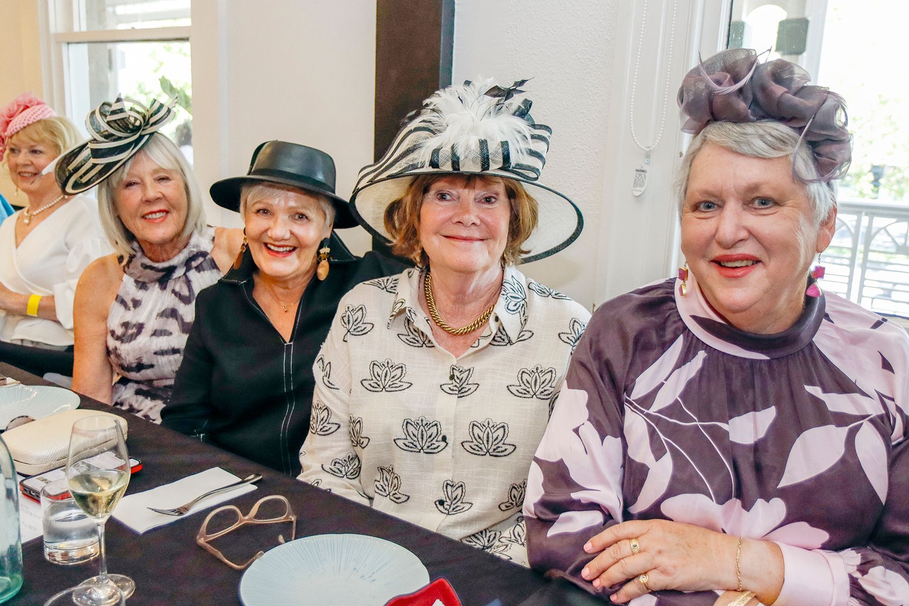 Women in elaborate hats at a luncheon. Smiling, indoors, wearing florals and other patterned outfits.
