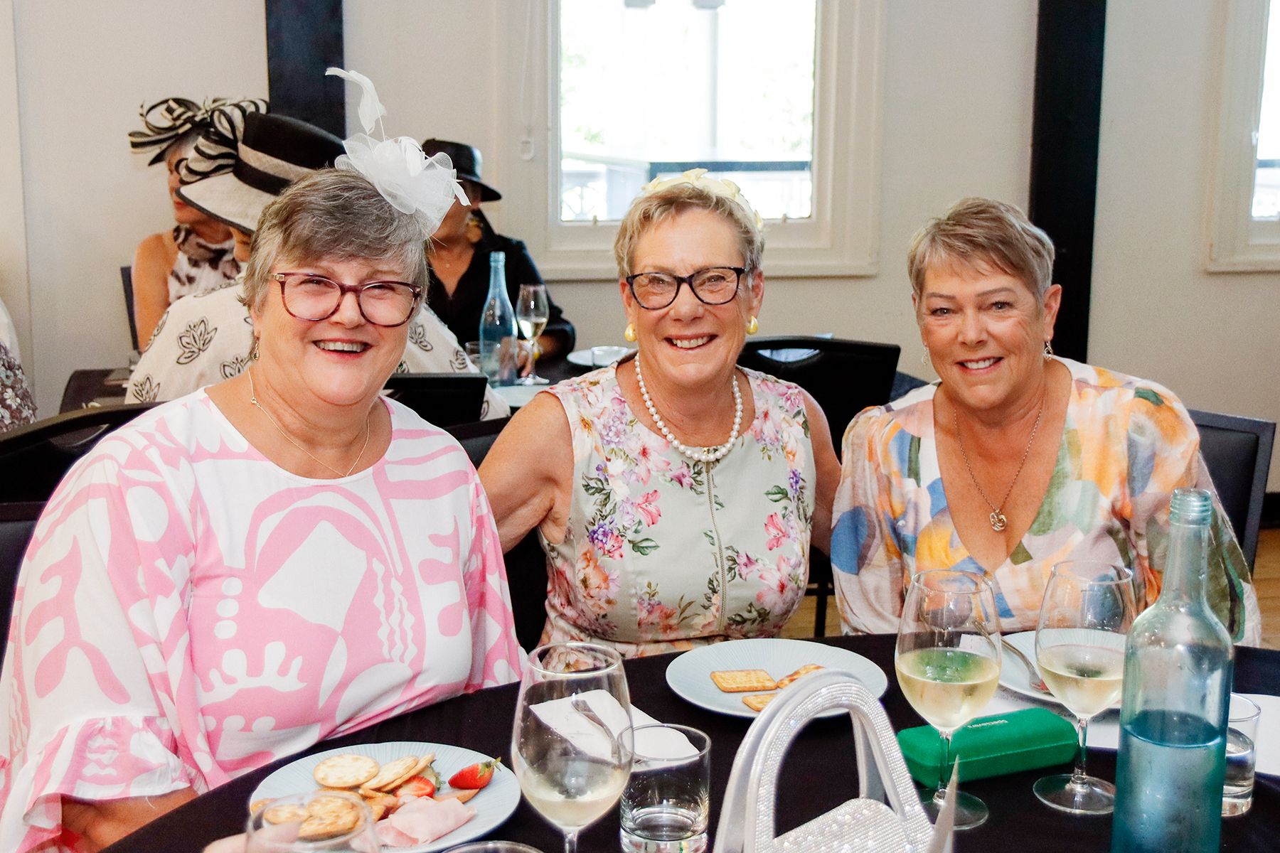 Three women at a table, smiling. One in pink, center in floral, right in patterned top. Glasses, snacks on table.