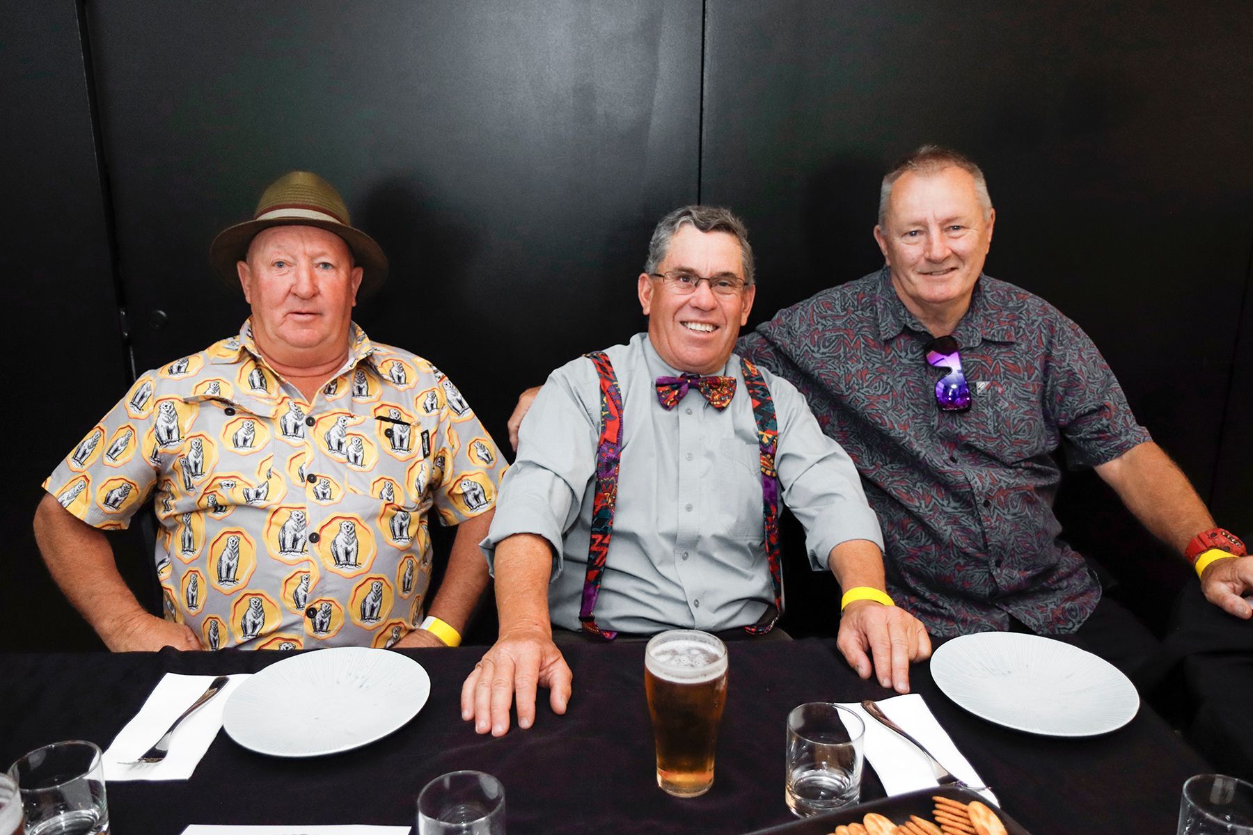 Three men at a table, one with a hat, another with a bow tie and suspenders. They sit with plates and drinks.