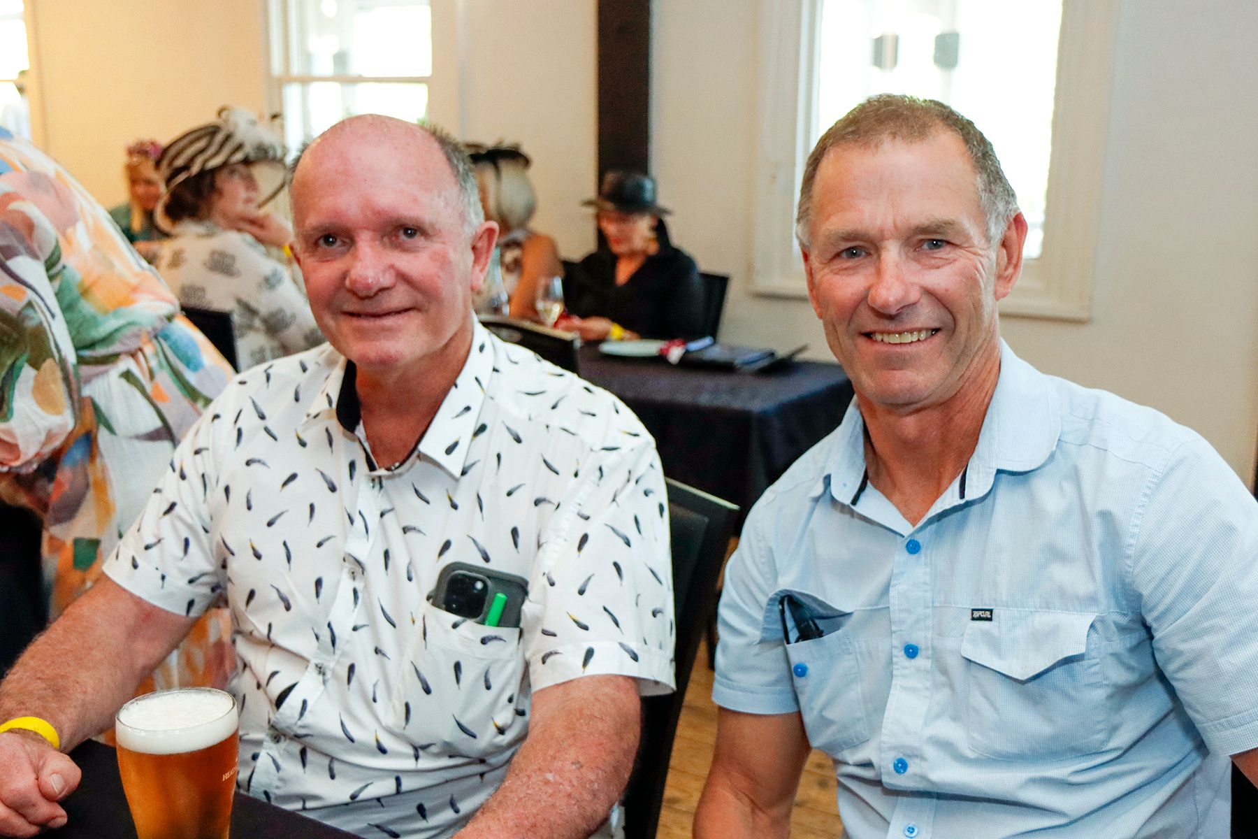 Two men smiling at a table; one wears a patterned shirt, the other a blue polo. They are indoors with others present.