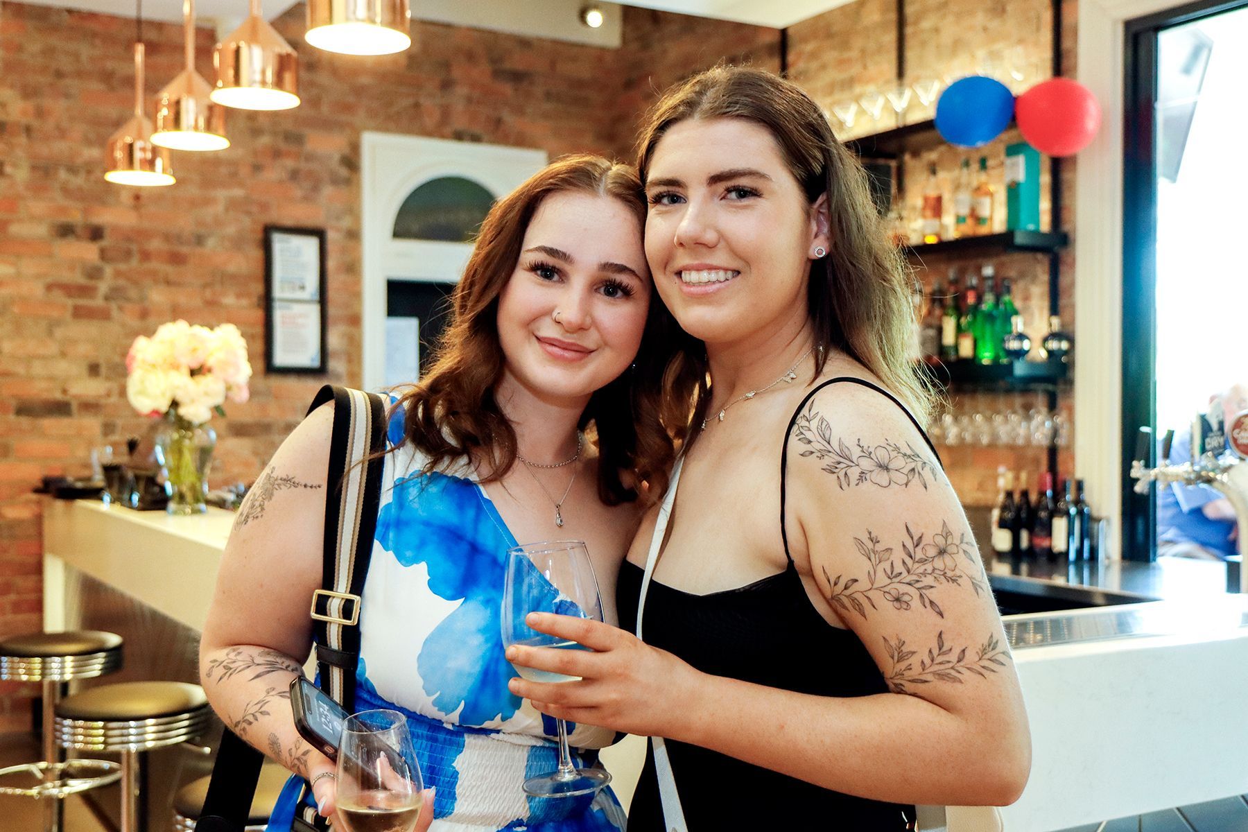 Two young women smiling at a bar, one in a blue dress, the other in black.