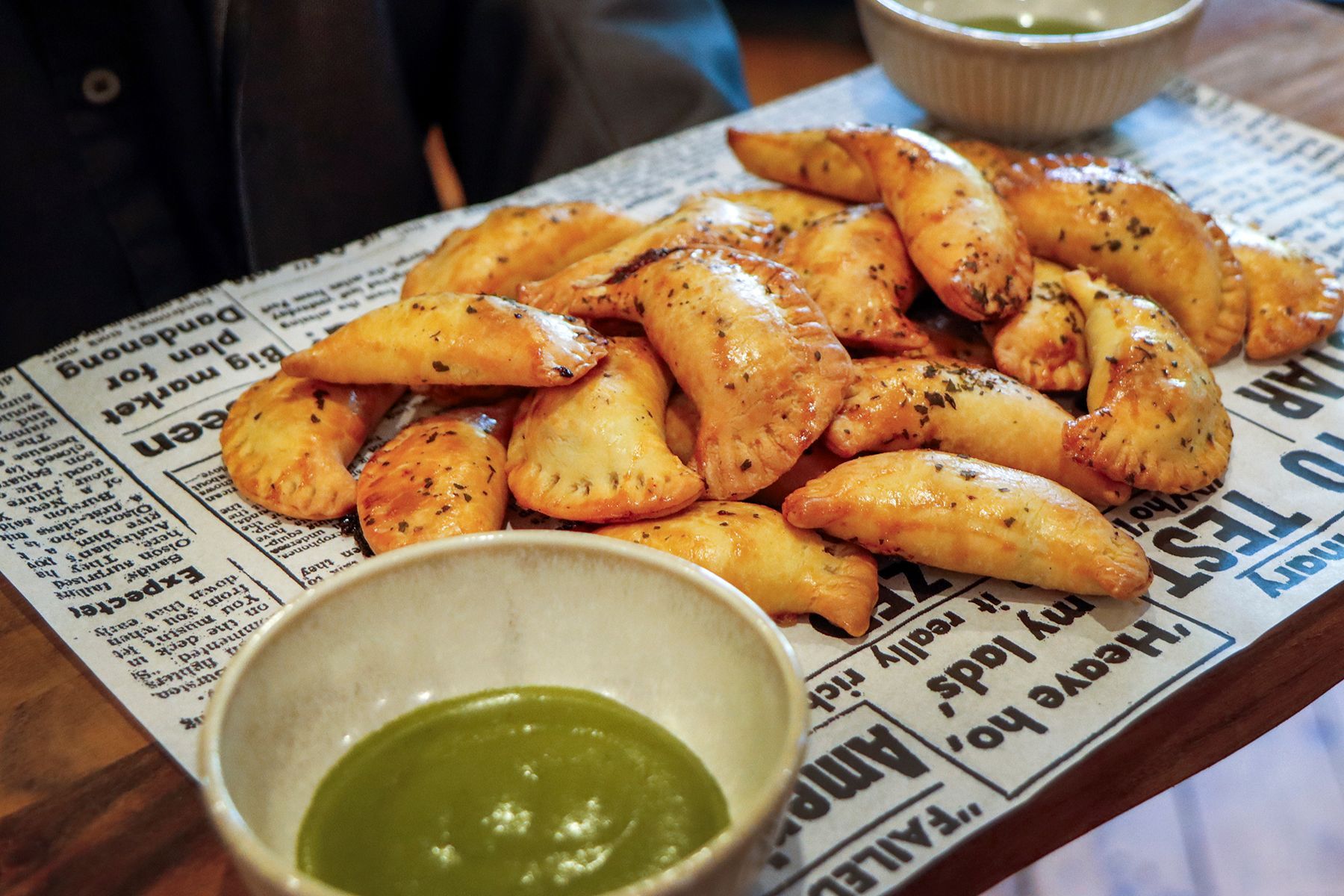 Golden-brown empanadas with green dipping sauce on newspaper-lined tray.