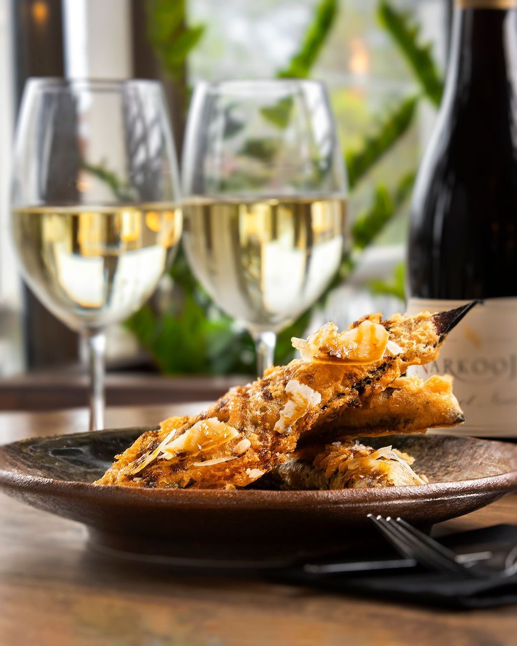 Two glasses of white wine, food on a plate, and a wine bottle on a wooden table.