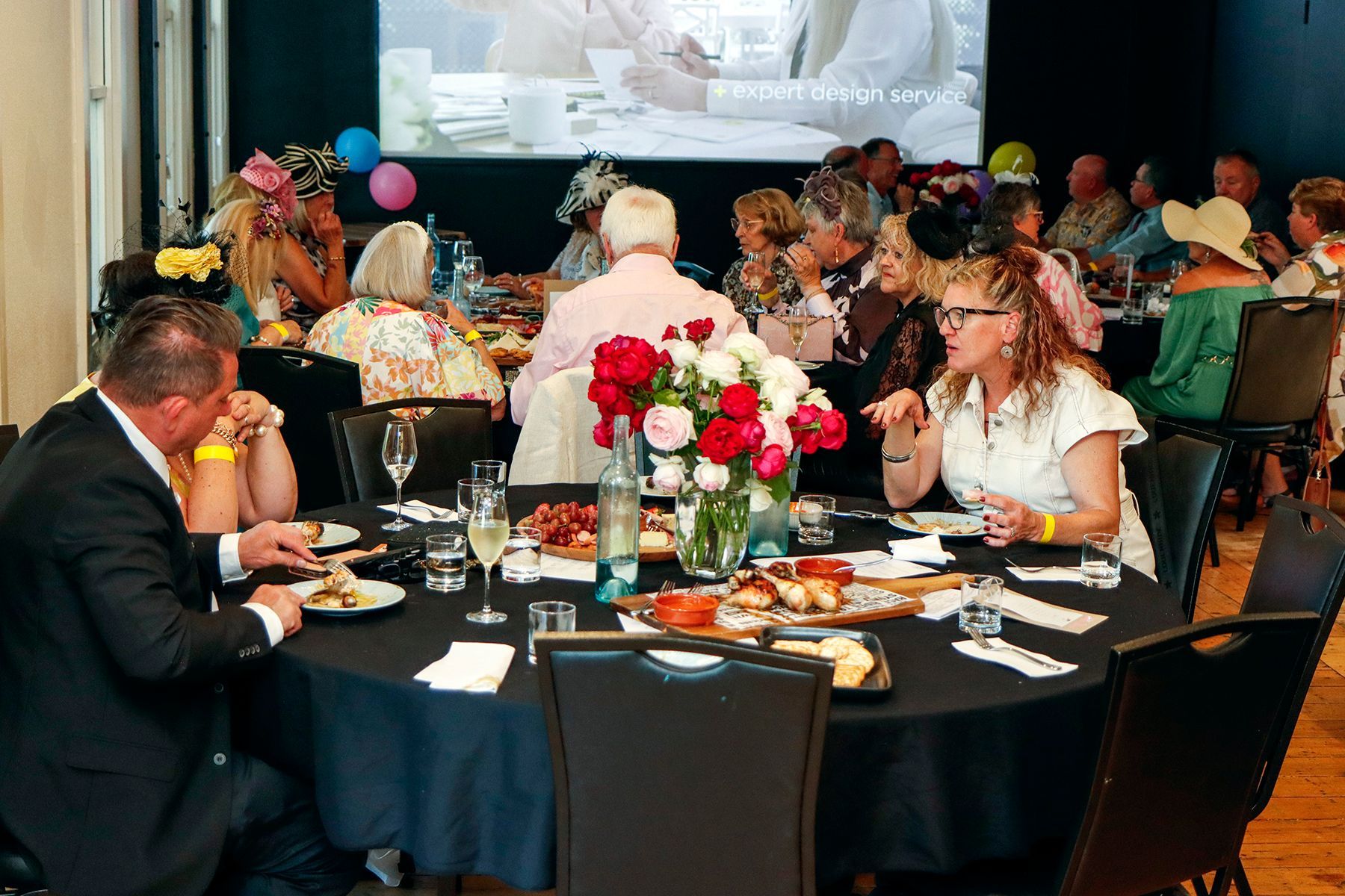 People at a formal event around tables, eating and socializing. Many wear hats, a projection screen visible.