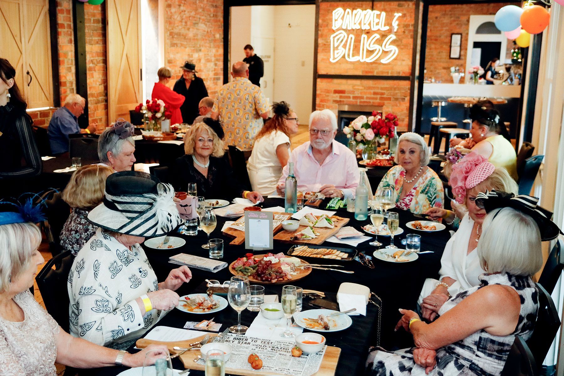 People in hats at a formal gathering around a table laden with food. 