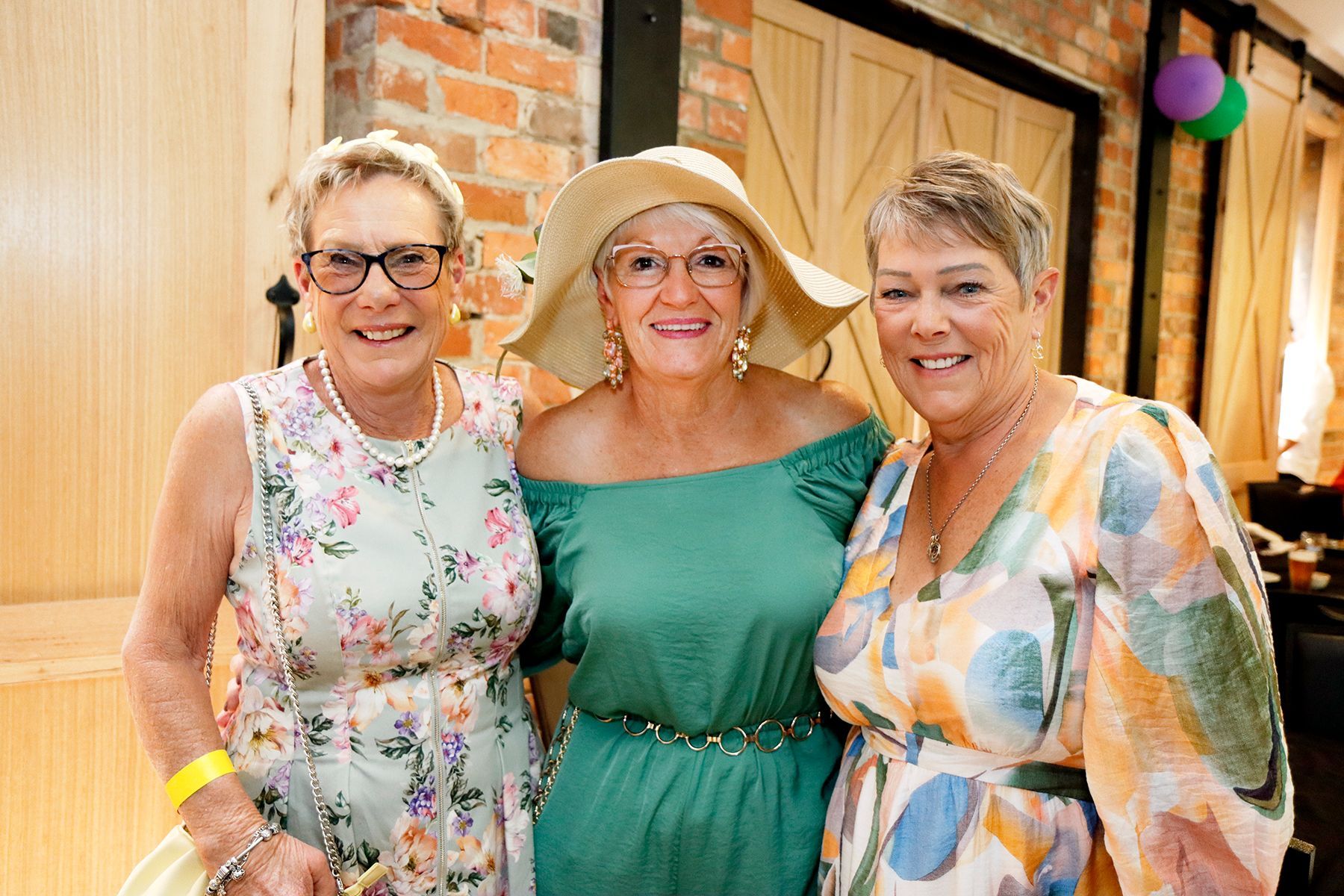 Three smiling women in dresses, one in a hat. Brick background, barn door.