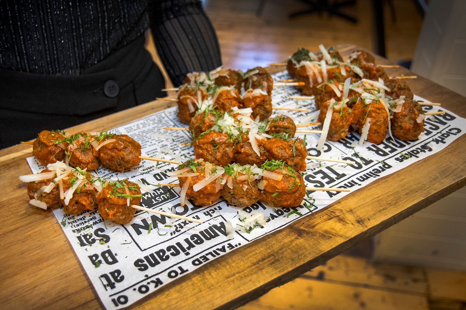 Meatballs on skewers, arranged on a wooden tray with newspaper, garnished with cheese and herbs.