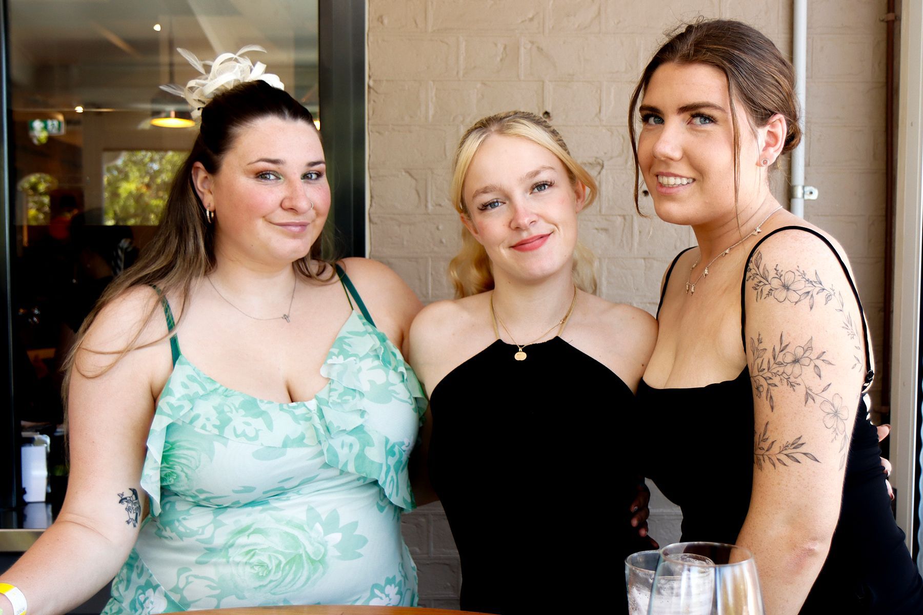 Three smiling women posing together outdoors. One wears green dress, center wears black halter top, other wears black dress, all looking at camera.