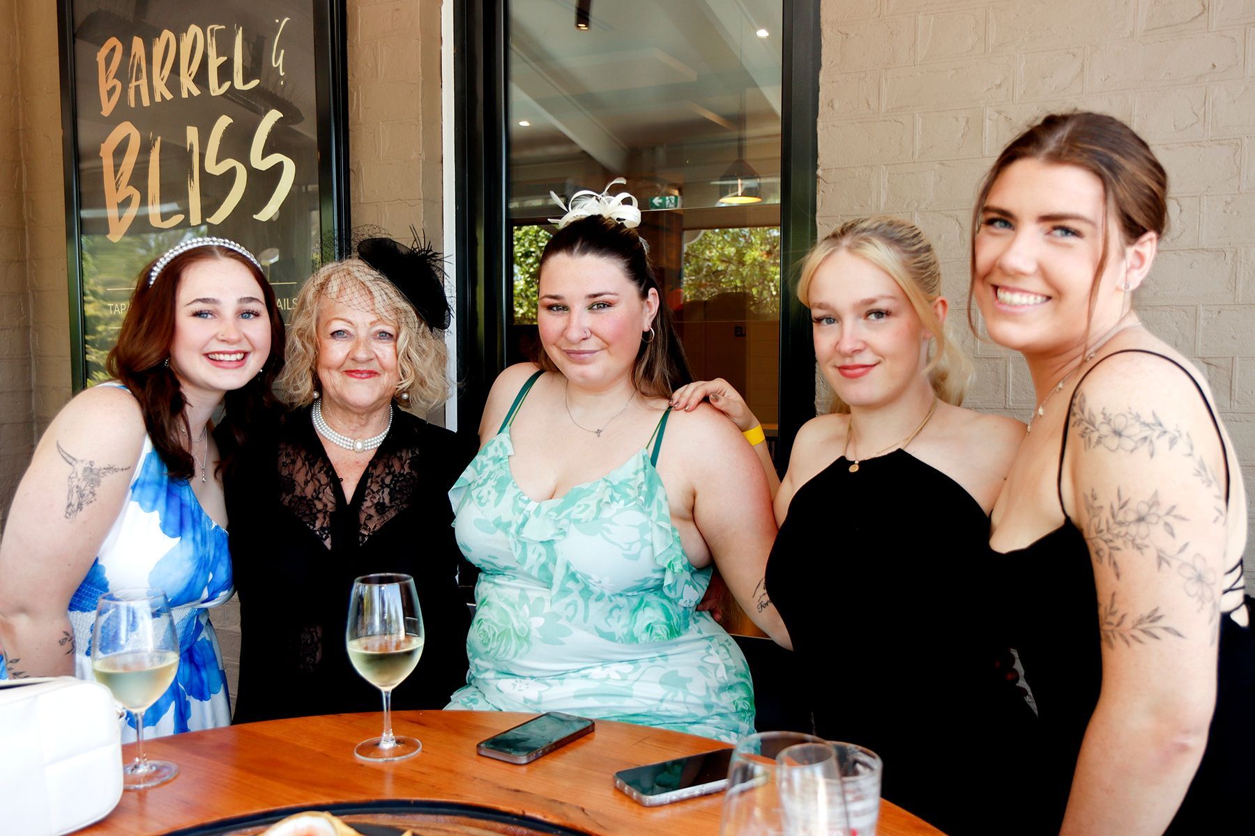 Five women smiling, posing near a table outdoors; one with a blue dress, a sign says 