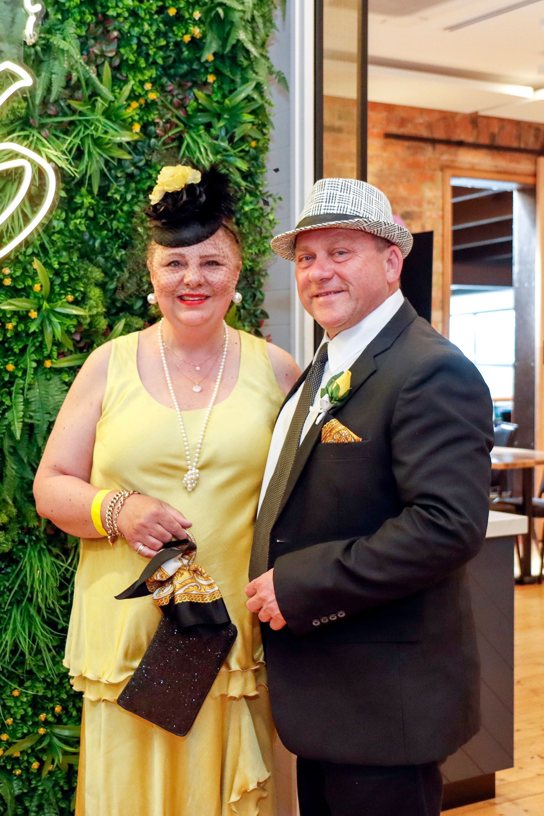 Couple in formal attire, woman in yellow dress and hat, man in suit and hat, near a green wall.