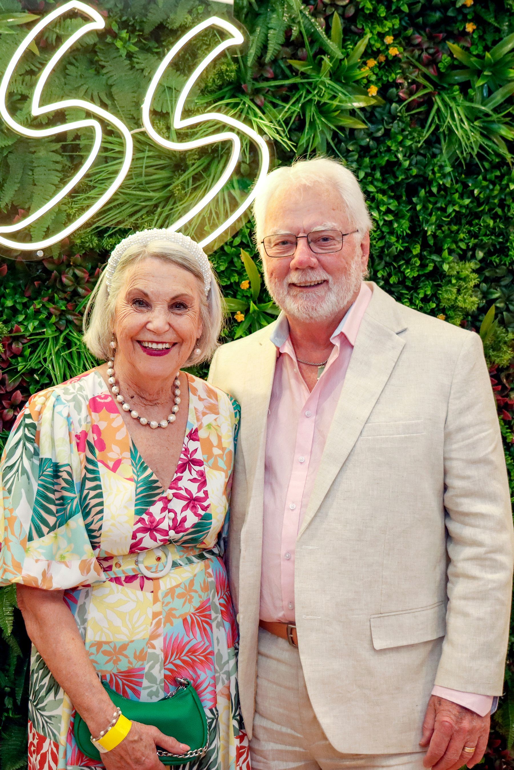 Couple smiling, posing in front of a leafy backdrop. Woman wears floral dress, man in light jacket, both in bright colors.