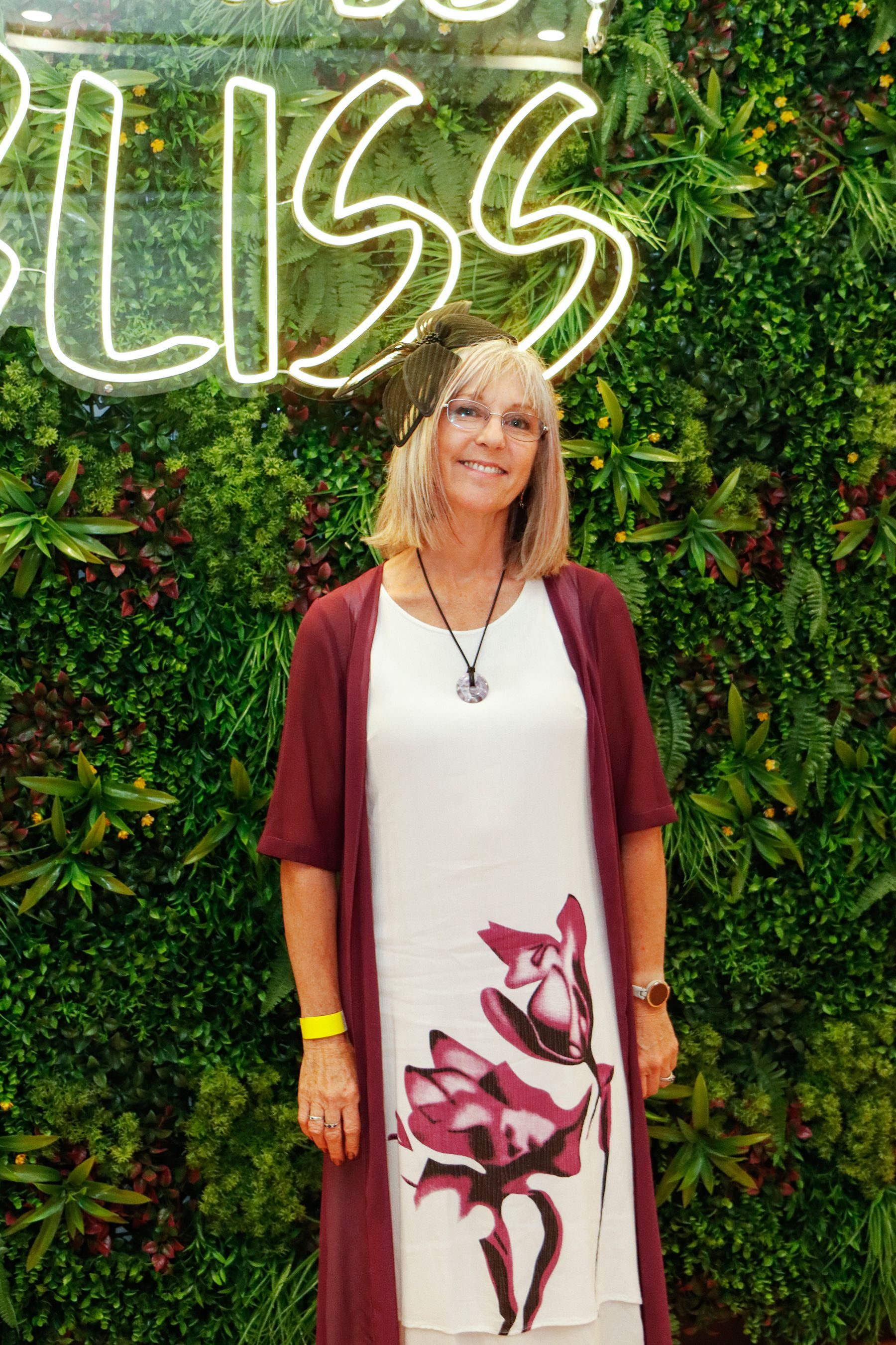 Woman in white dress with floral print and maroon cardigan stands in front of a greenery wall with neon 