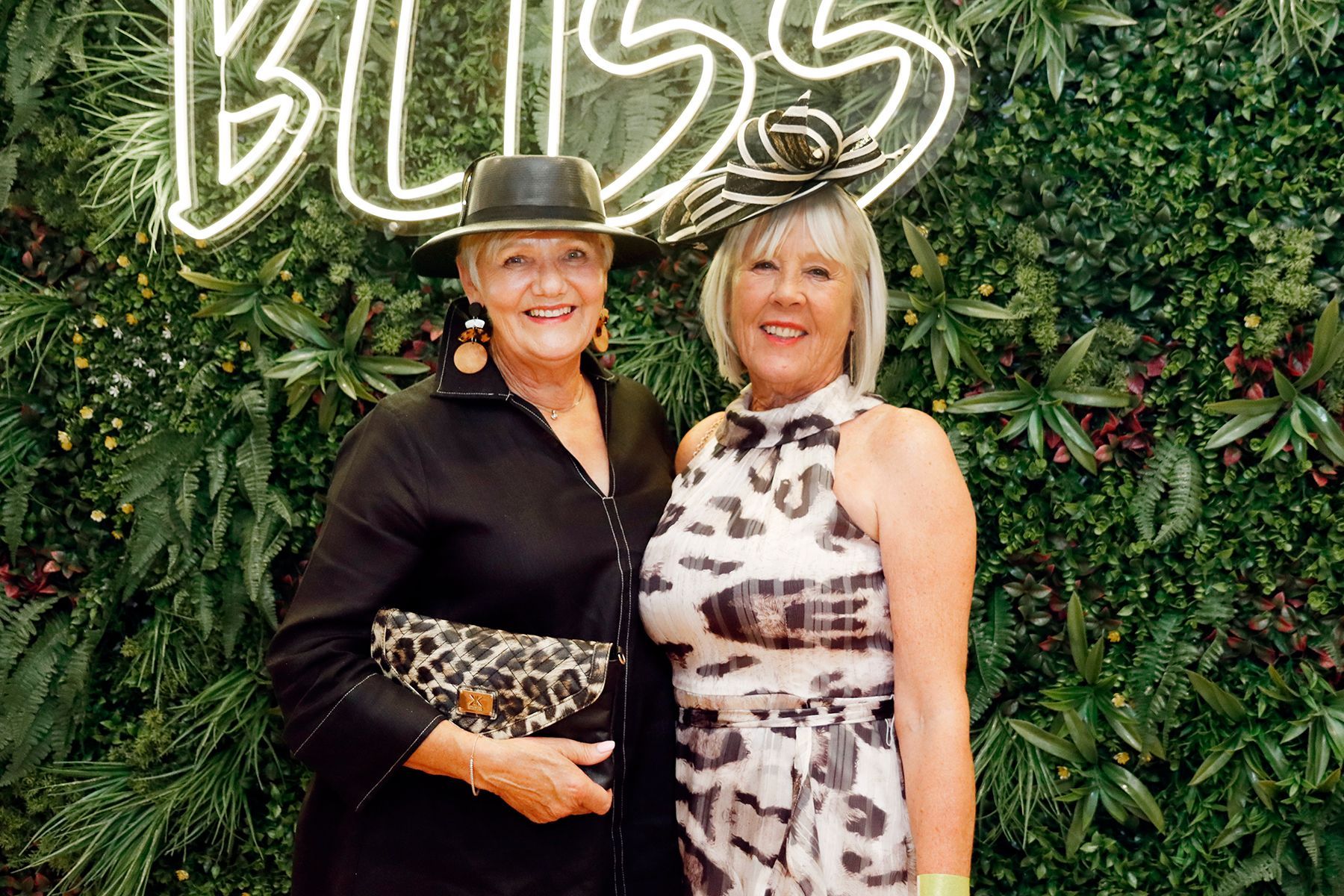 Two women in hats pose in front of a green floral wall with 