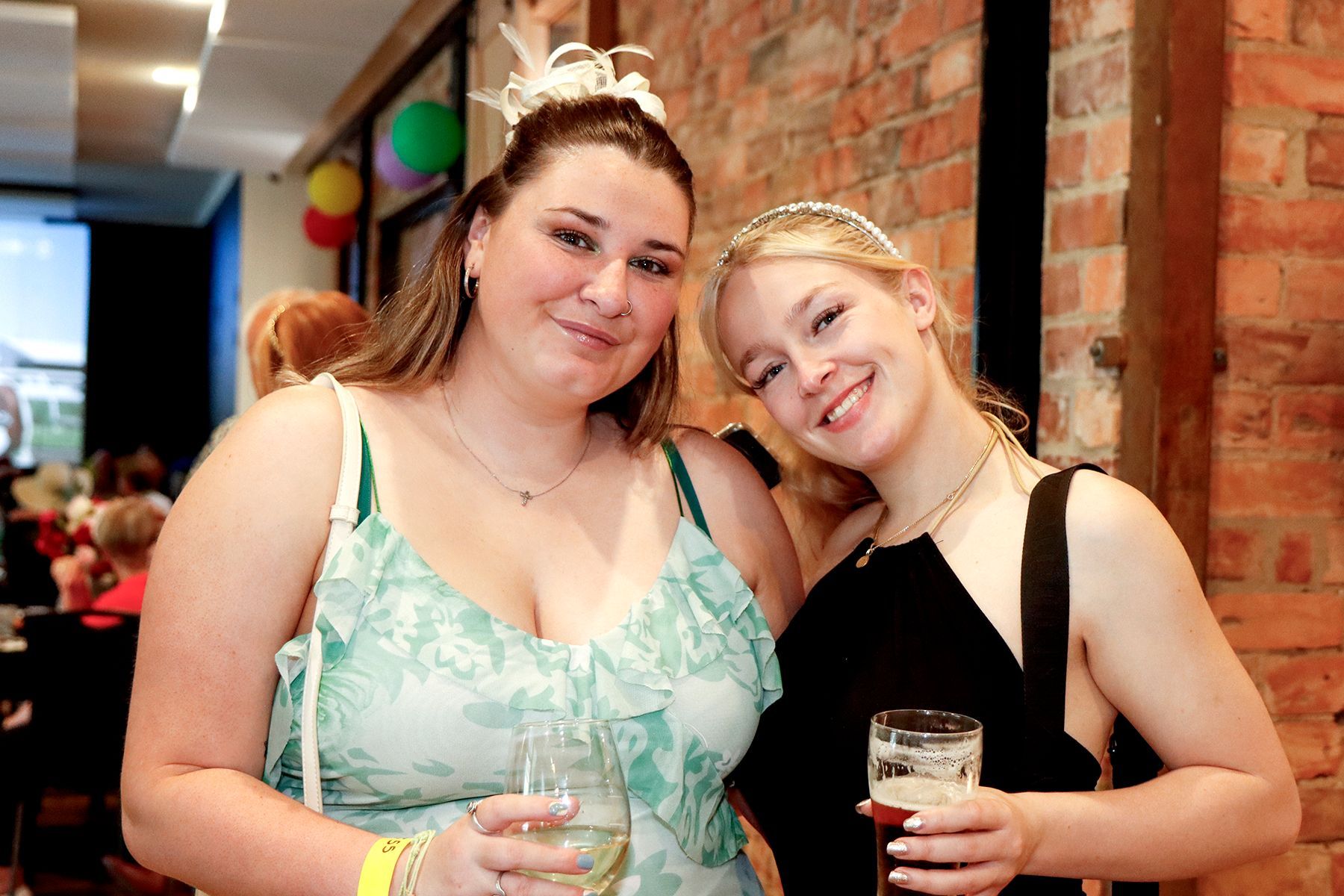 Two women smiling, one with a drink, near a brick wall. Blonde and brunette wearing dresses.
