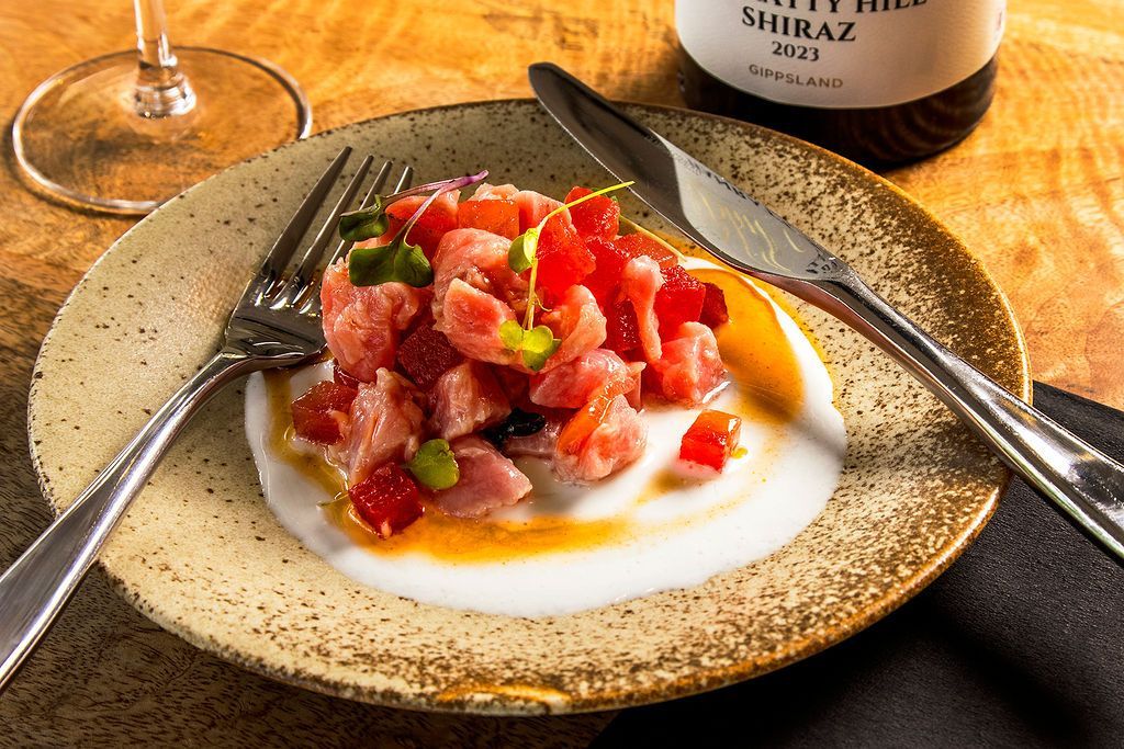 Plate of diced red tuna with white sauce, fork, knife, wine glass, and bottle of Shiraz.