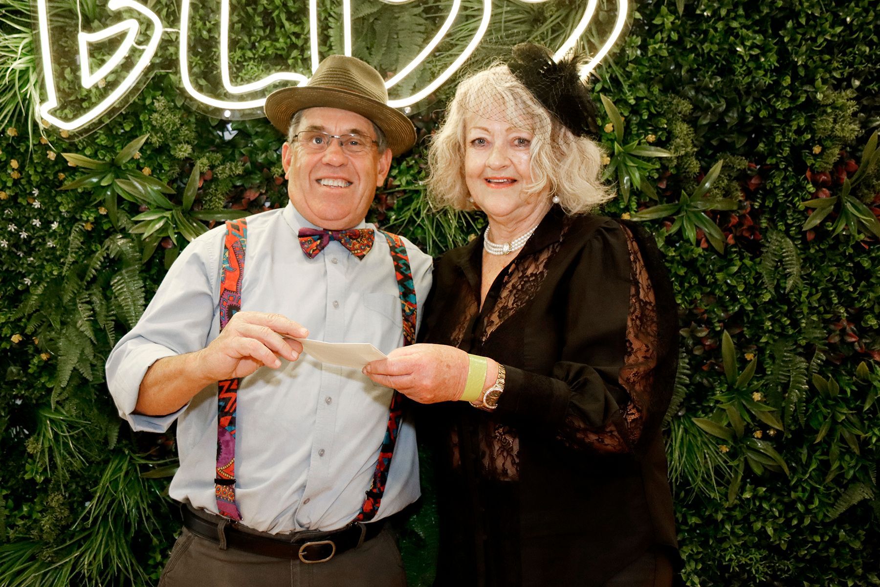 Man in hat and suspenders gives check to woman in black dress; green plant wall backdrop.