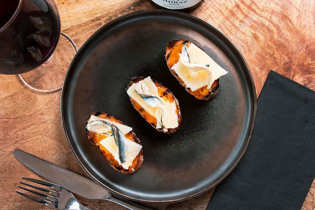 Three bruschetta appetizers topped with cheese and anchovies on a dark plate, next to wine and silverware.