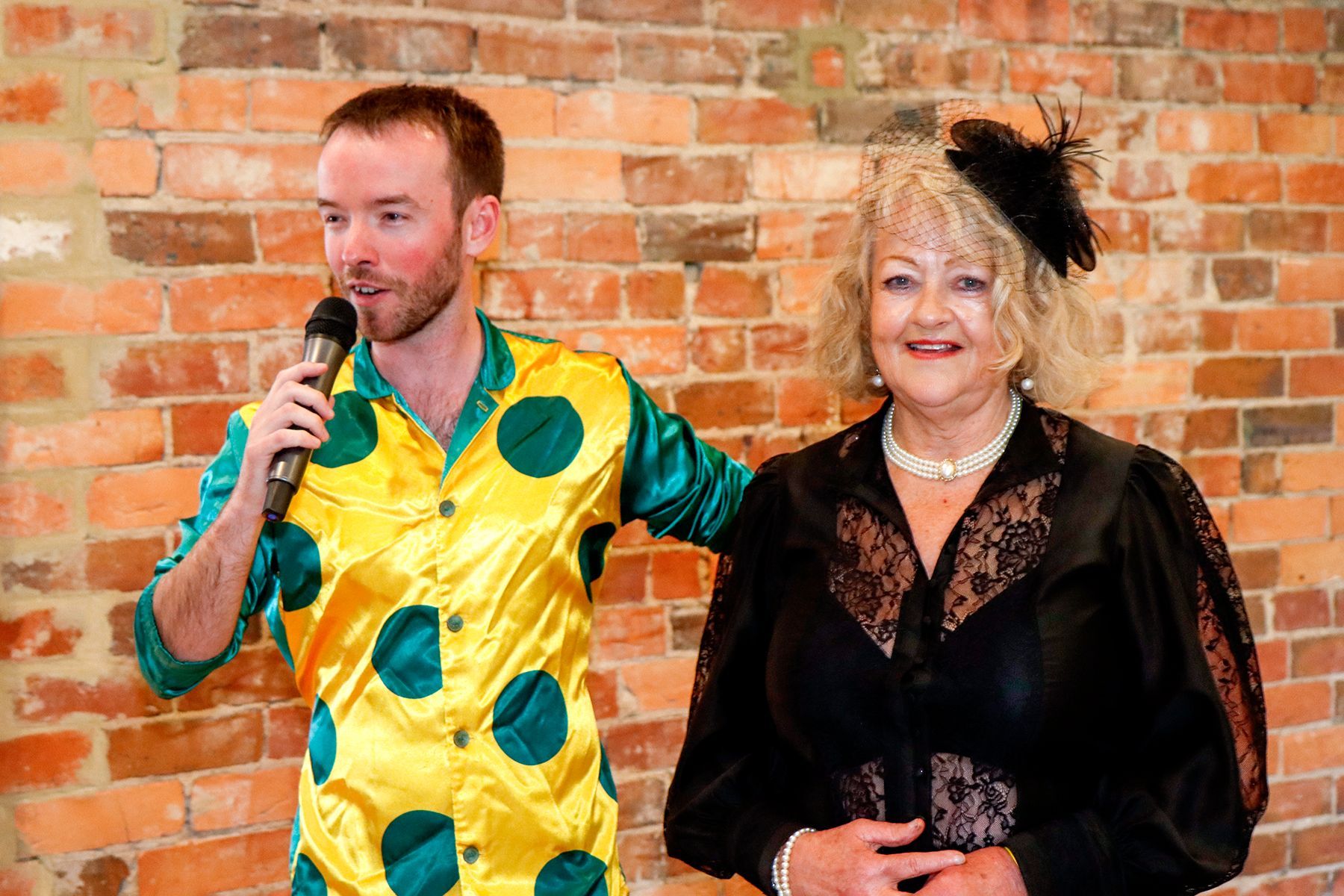 Man in a yellow and green polka-dot shirt with a microphone next to a woman in a black dress, by a brick wall.