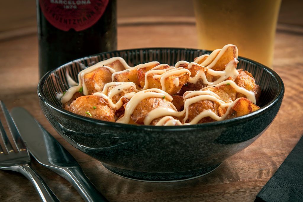Bowl of fried potatoes drizzled with sauce, on a wooden table with a bottle and cutlery.