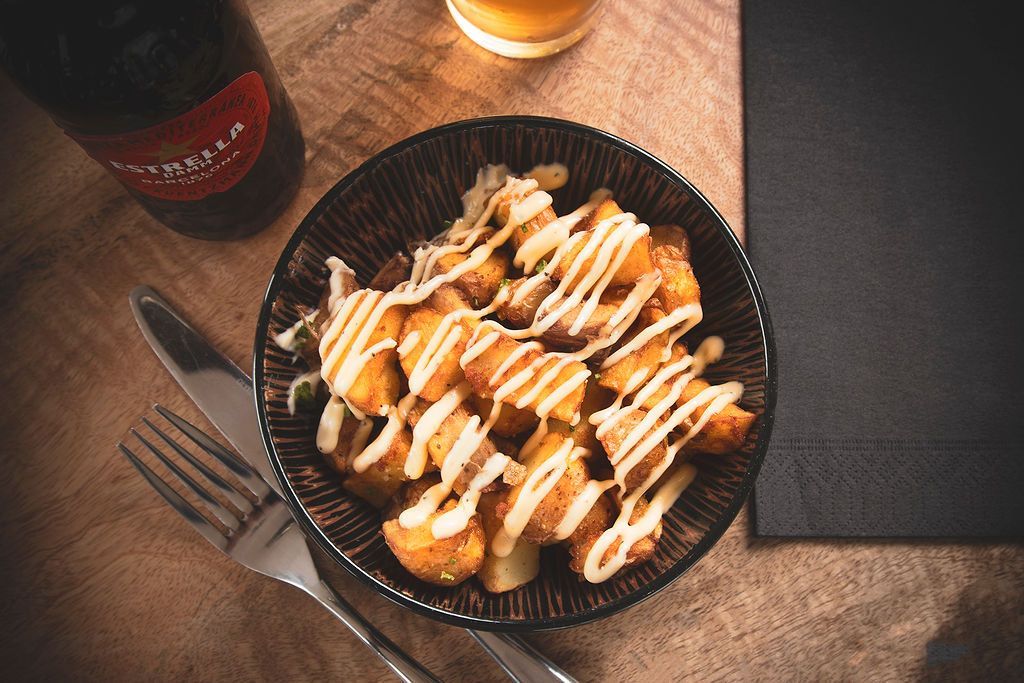 Bowl of fried food drizzled with sauce on a wooden table, with a bottle, glass, fork, knife, and menu.