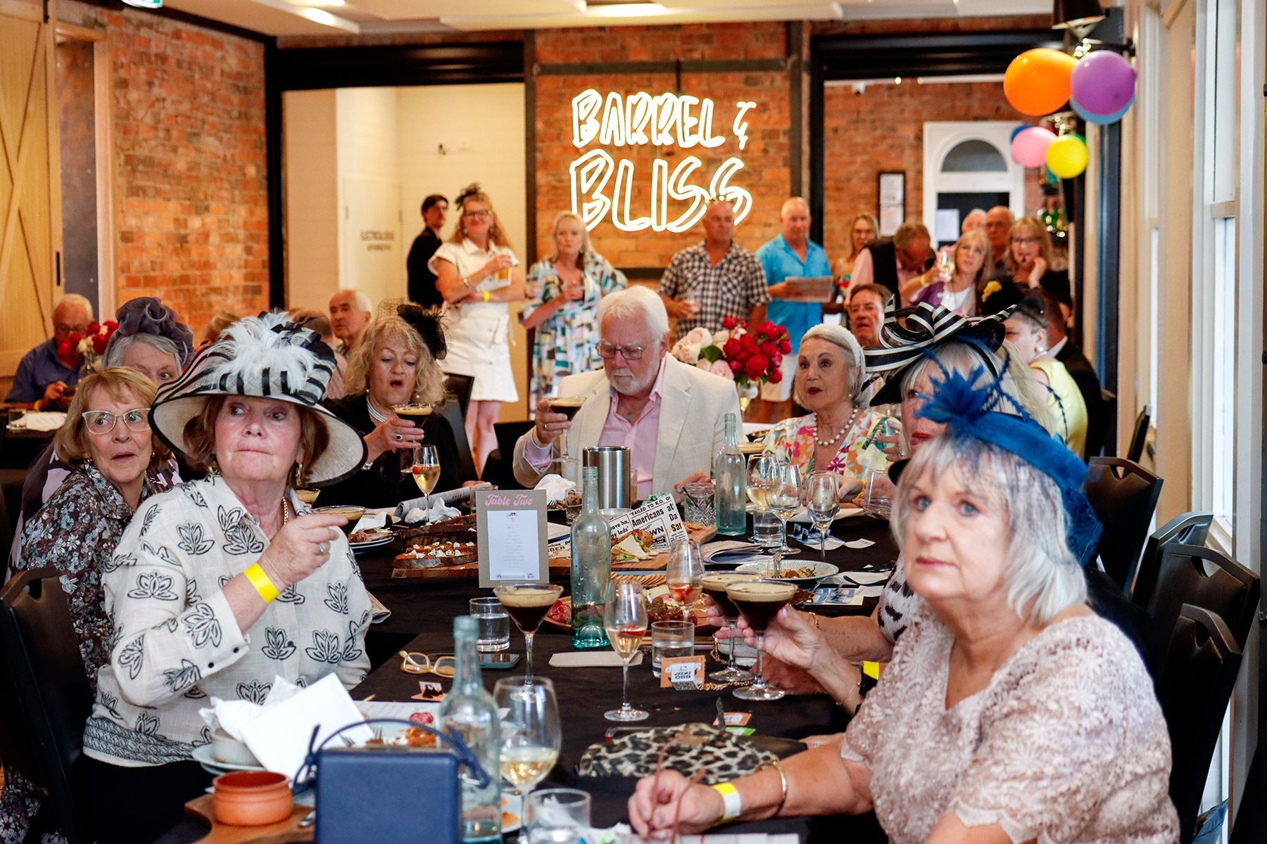 People in hats and formal attire at a luncheon, many smiling and raising glasses, inside a restaurant.