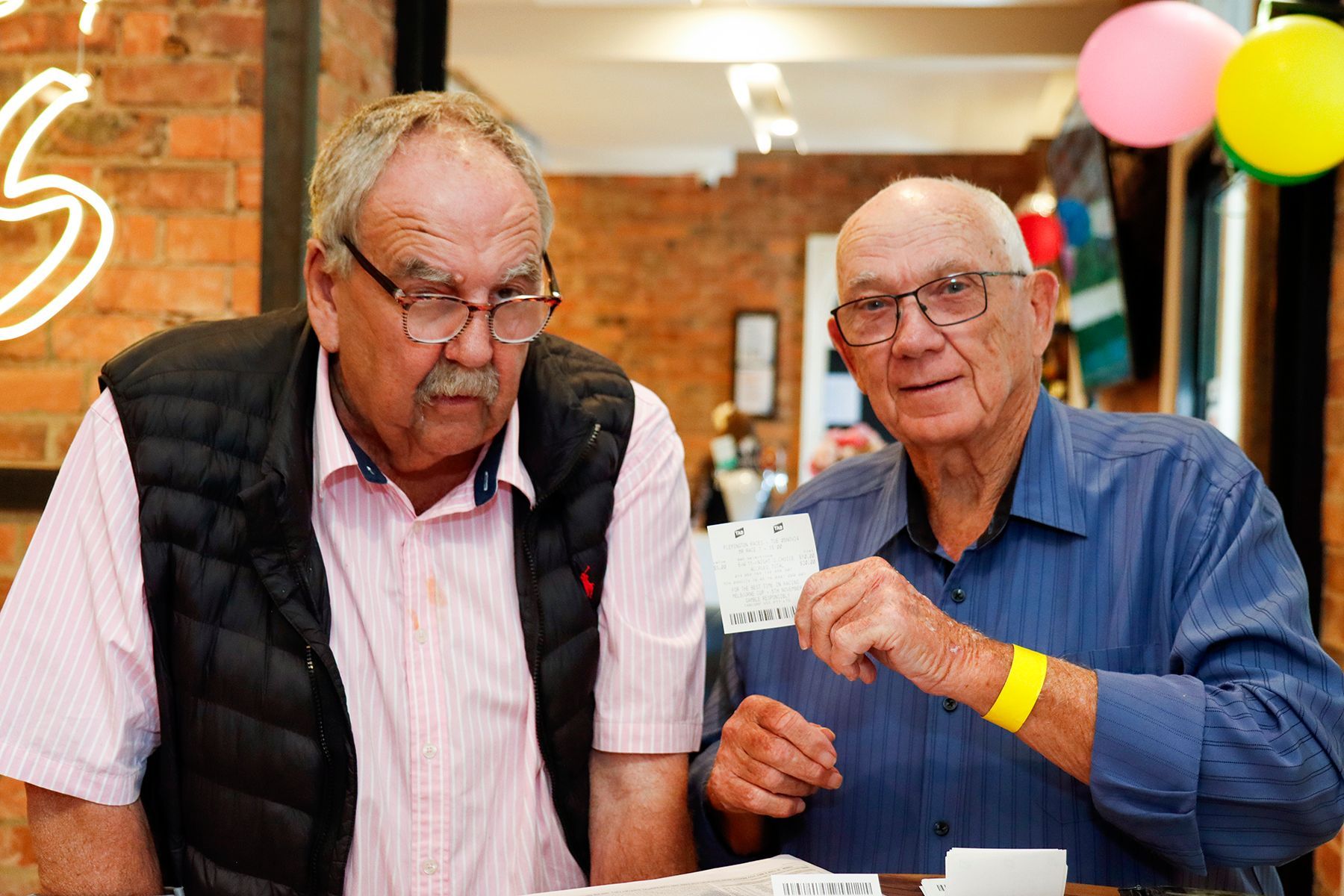 Two older men at a table. One holds a card, the other looks on. Indoor setting with brick wall.