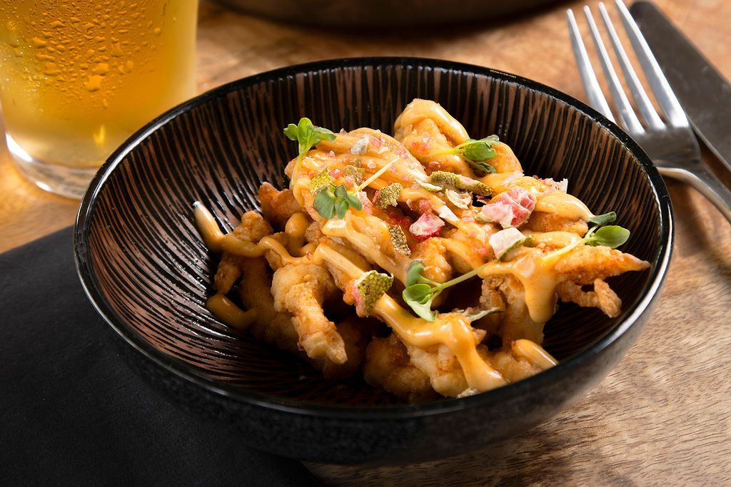 Fried calamari in a black bowl, topped with sauce and garnishes. A beer and silverware are in the background.