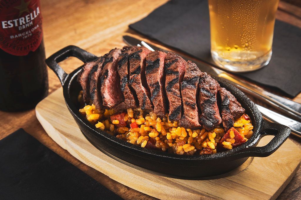 Grilled steak over rice in a black skillet with a beer and a bottle of Estrella beer on a wooden table.