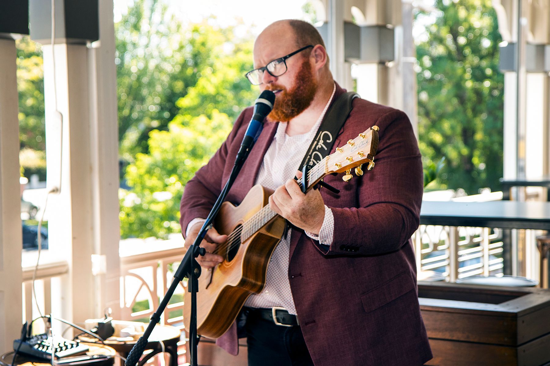 Man with beard sings and plays acoustic guitar outdoors, wearing burgundy jacket.