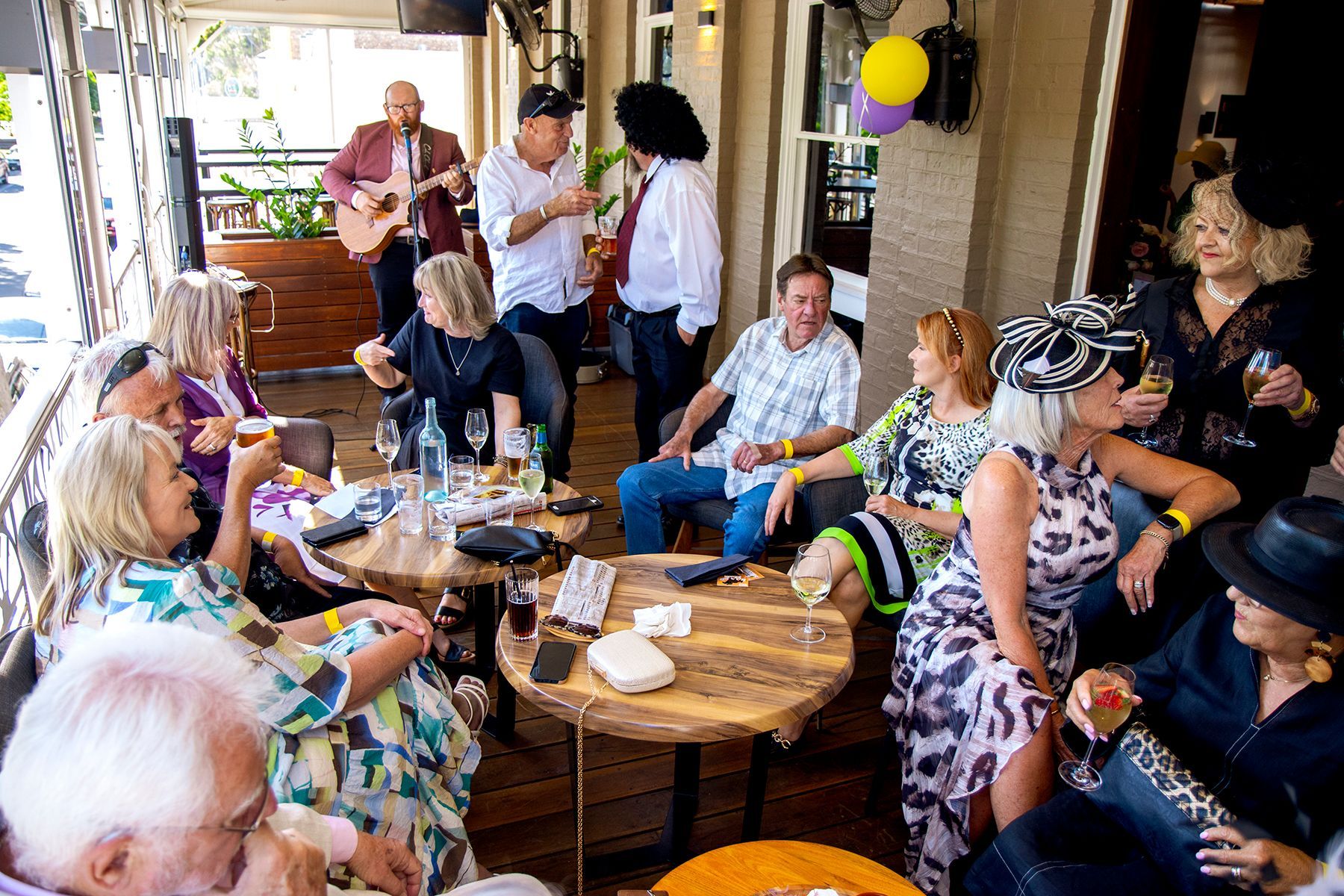 People socializing outdoors at a venue with live music; various ages, colorful attire, relaxed expressions.