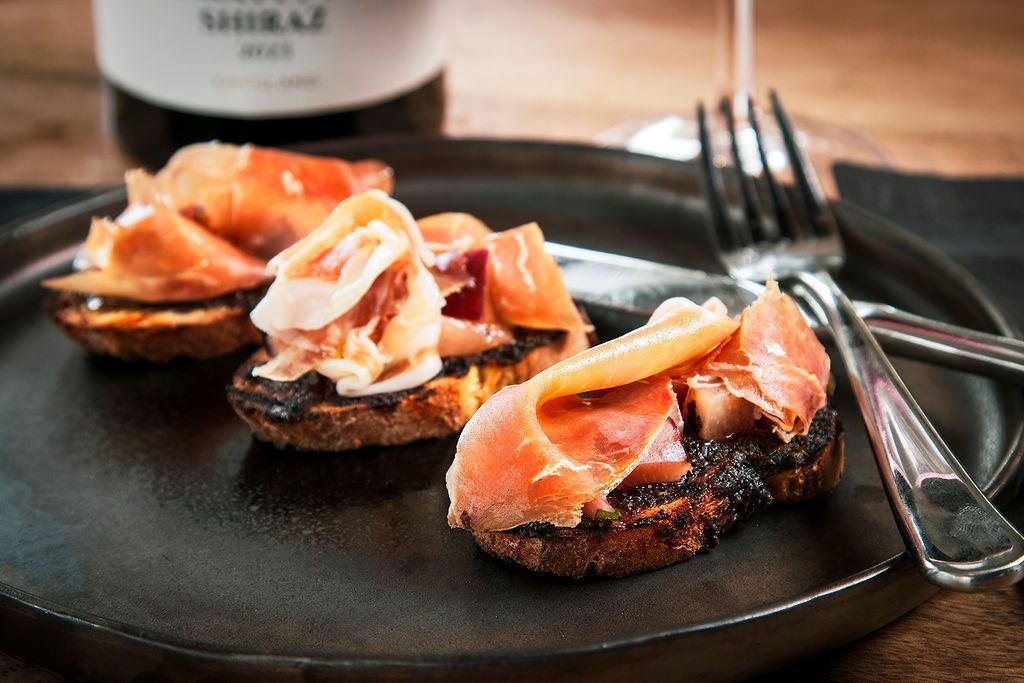 Prosciutto on toasted bread slices, served on a black plate with a fork and wine.