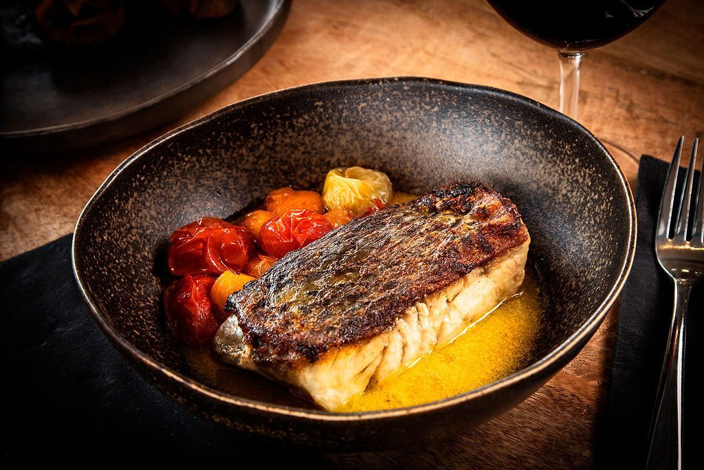 Seared fish fillet in a bowl with tomatoes, yellow sauce. Fork and wine glass in background.