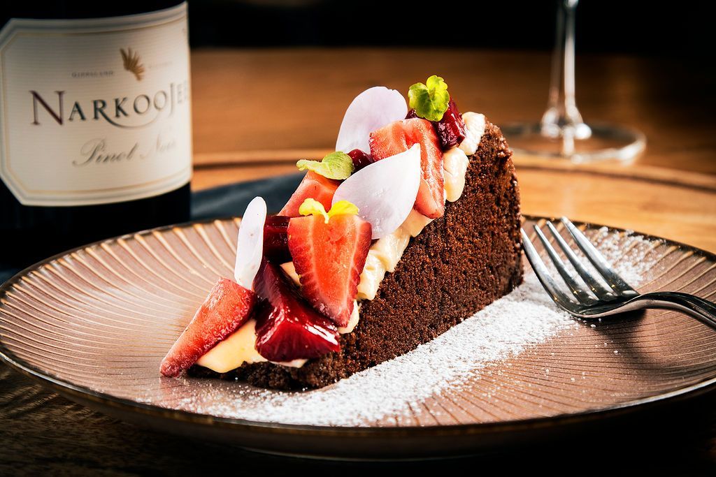 Slice of chocolate cake with strawberries and powdered sugar on a plate, wine bottle in the background.
