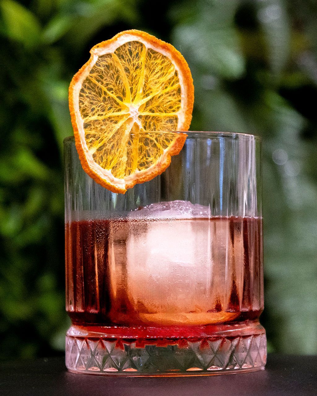 Cocktail in a crystal glass with orange slice garnish and ice. Set against blurred green foliage.