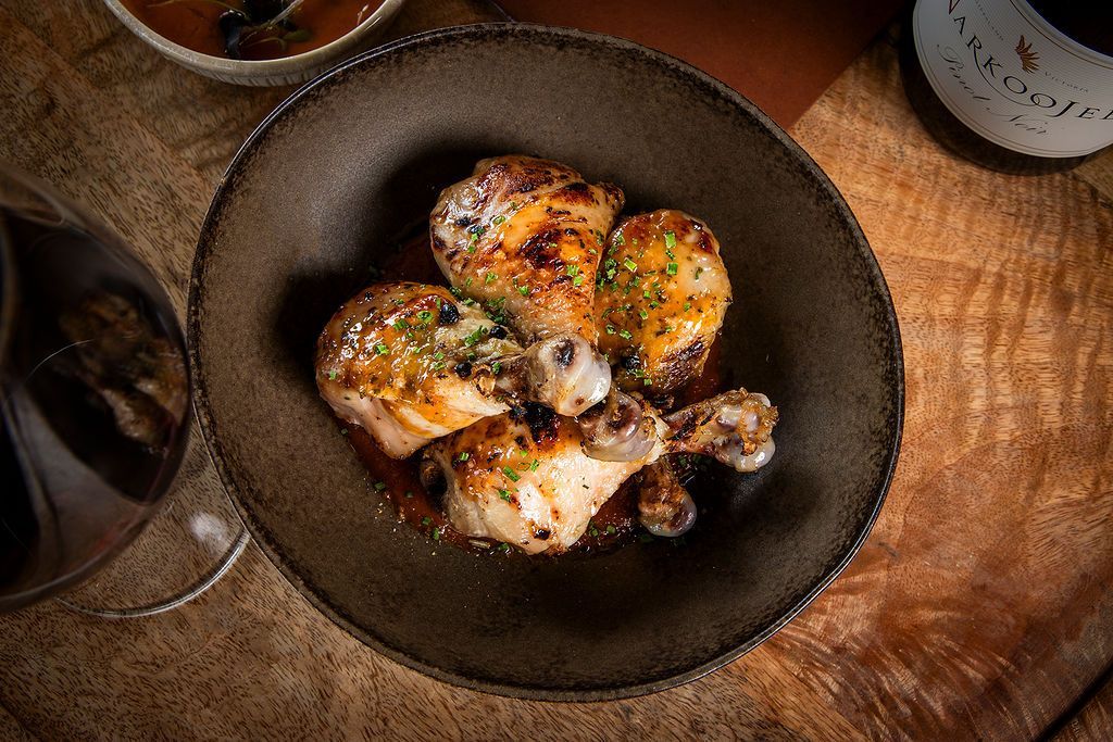 Chicken drumsticks in a dark bowl, with a glass of red wine and sauce on a wooden surface.