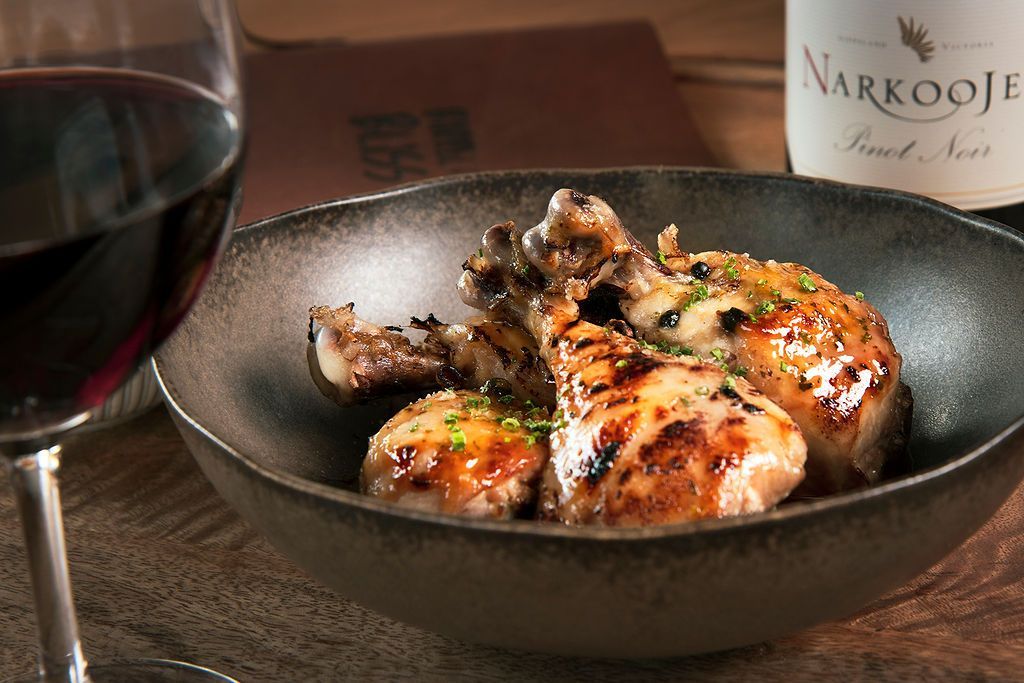 Roasted chicken in a dark bowl with a glass of red wine and a bottle of wine in the background.