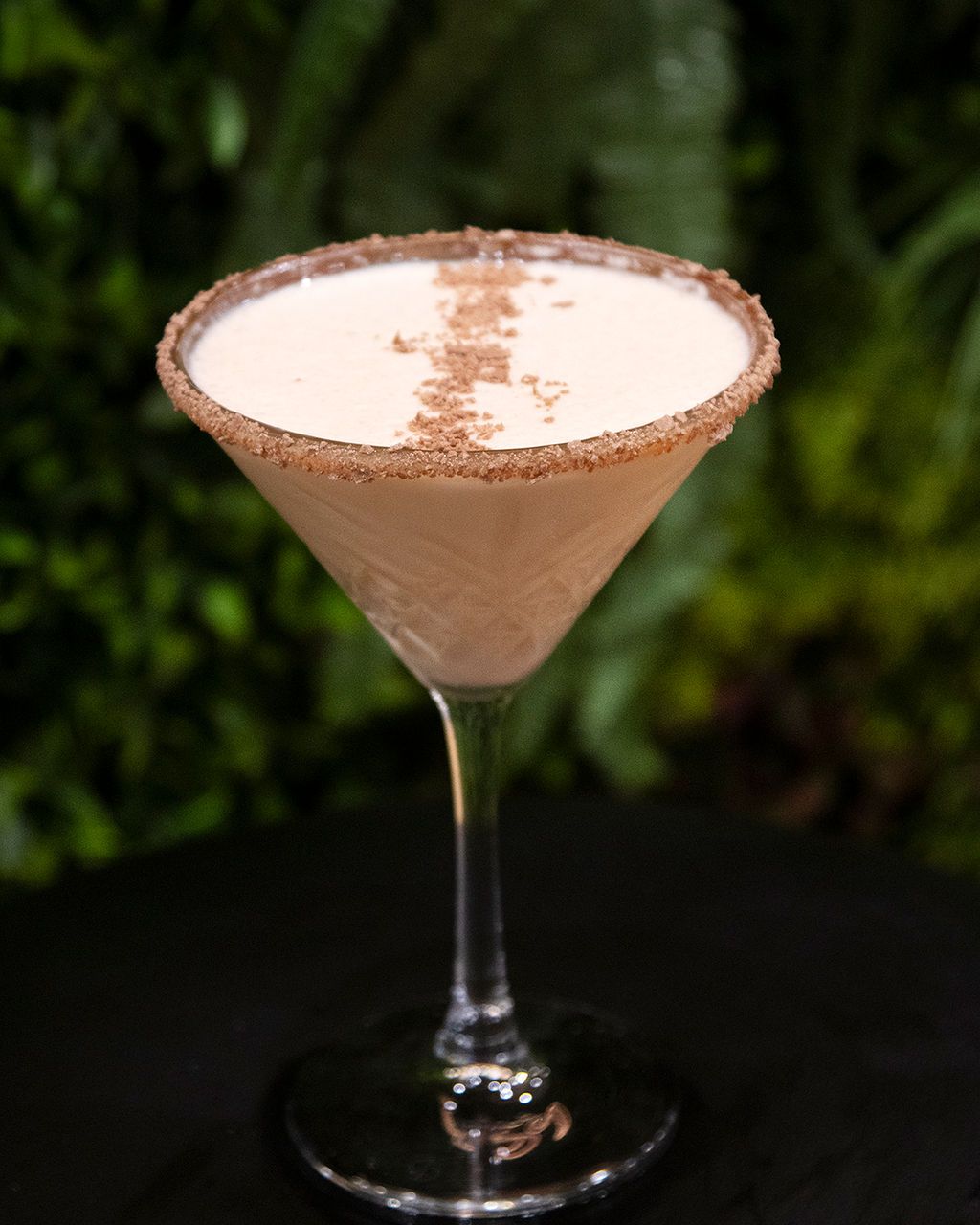 Cocktail in a martini glass with a cinnamon sugar rim, on a black surface, against a green backdrop.