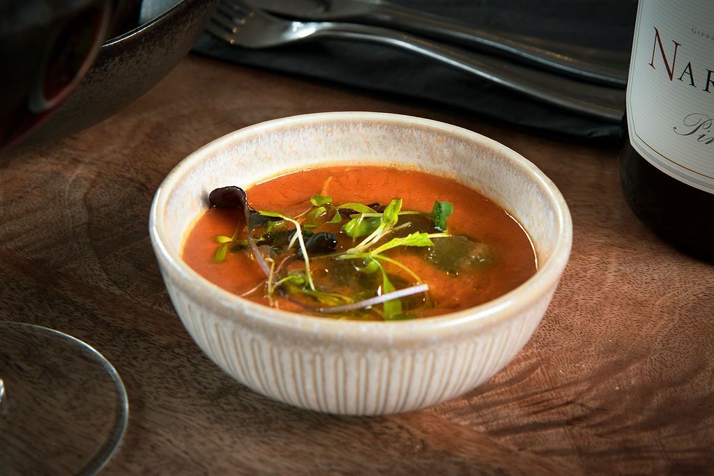 Bowl of red soup with microgreens on a wooden table; silverware and wine bottle in background.