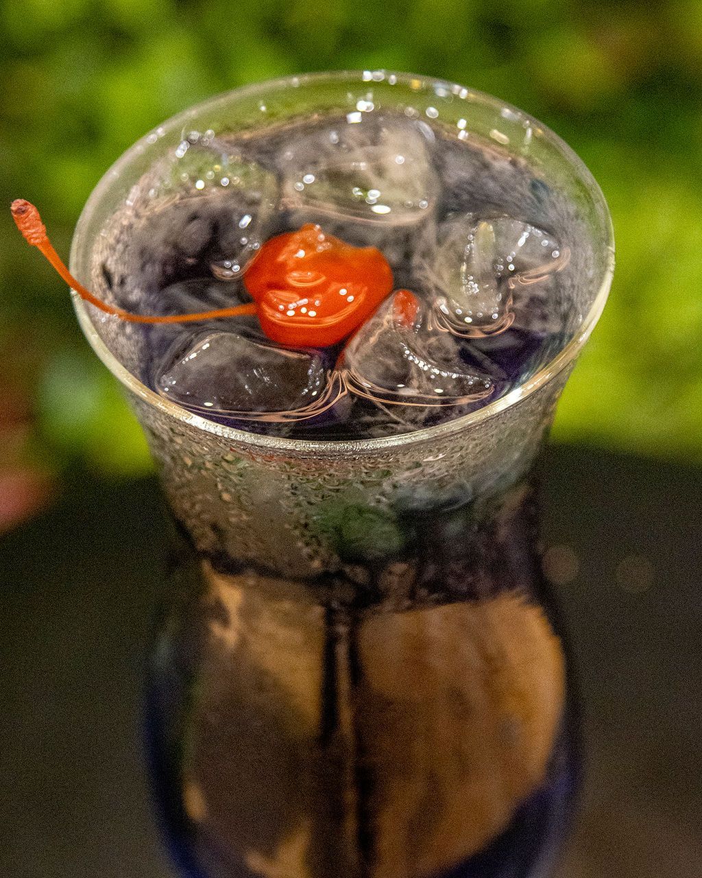 Cocktail with ice and a maraschino cherry, in a blue-tinted glass, reflecting on a dark surface.